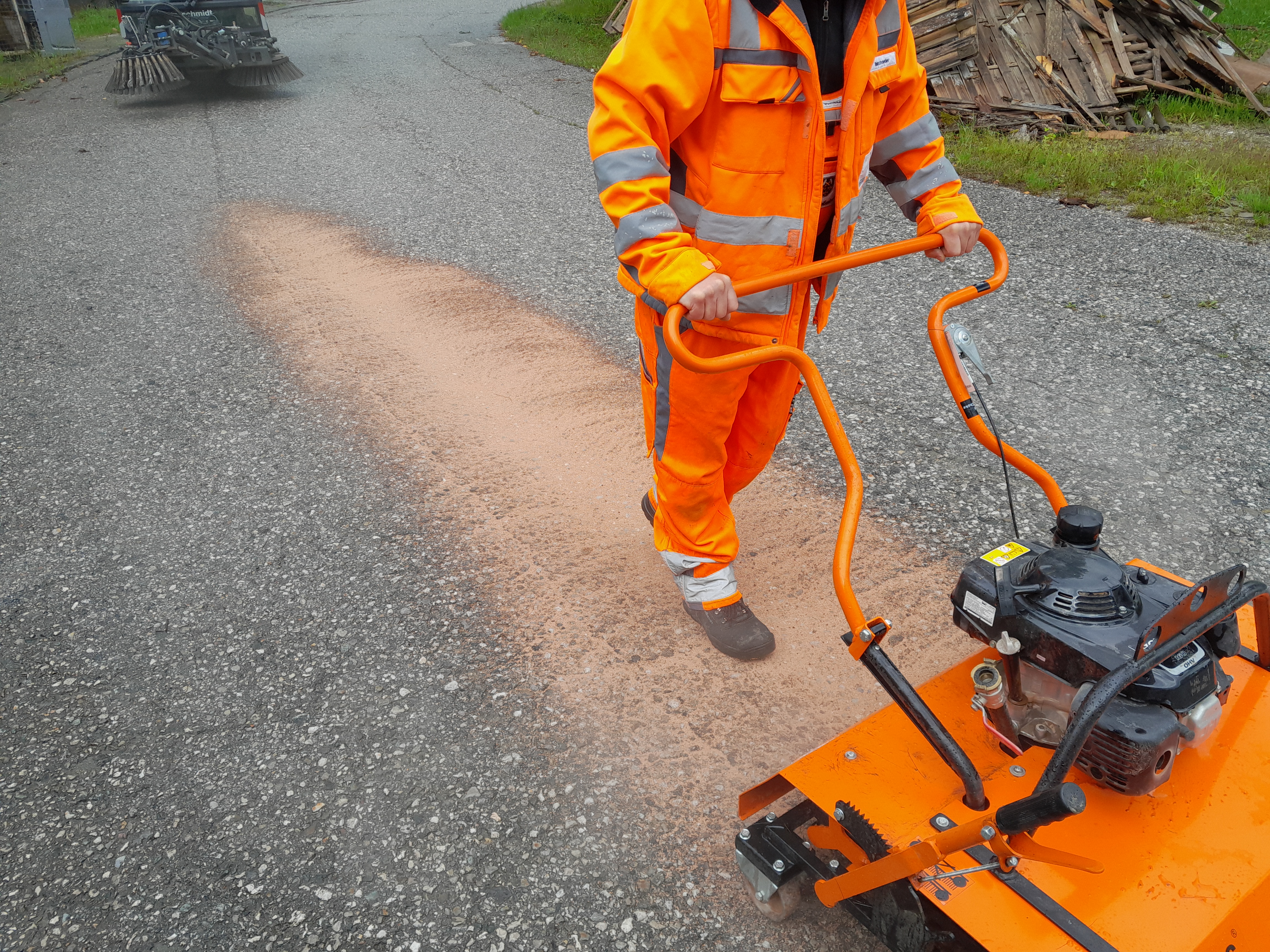 A worker in an orange safety suit operates a machine spreading material on a gravel road – Optimas.