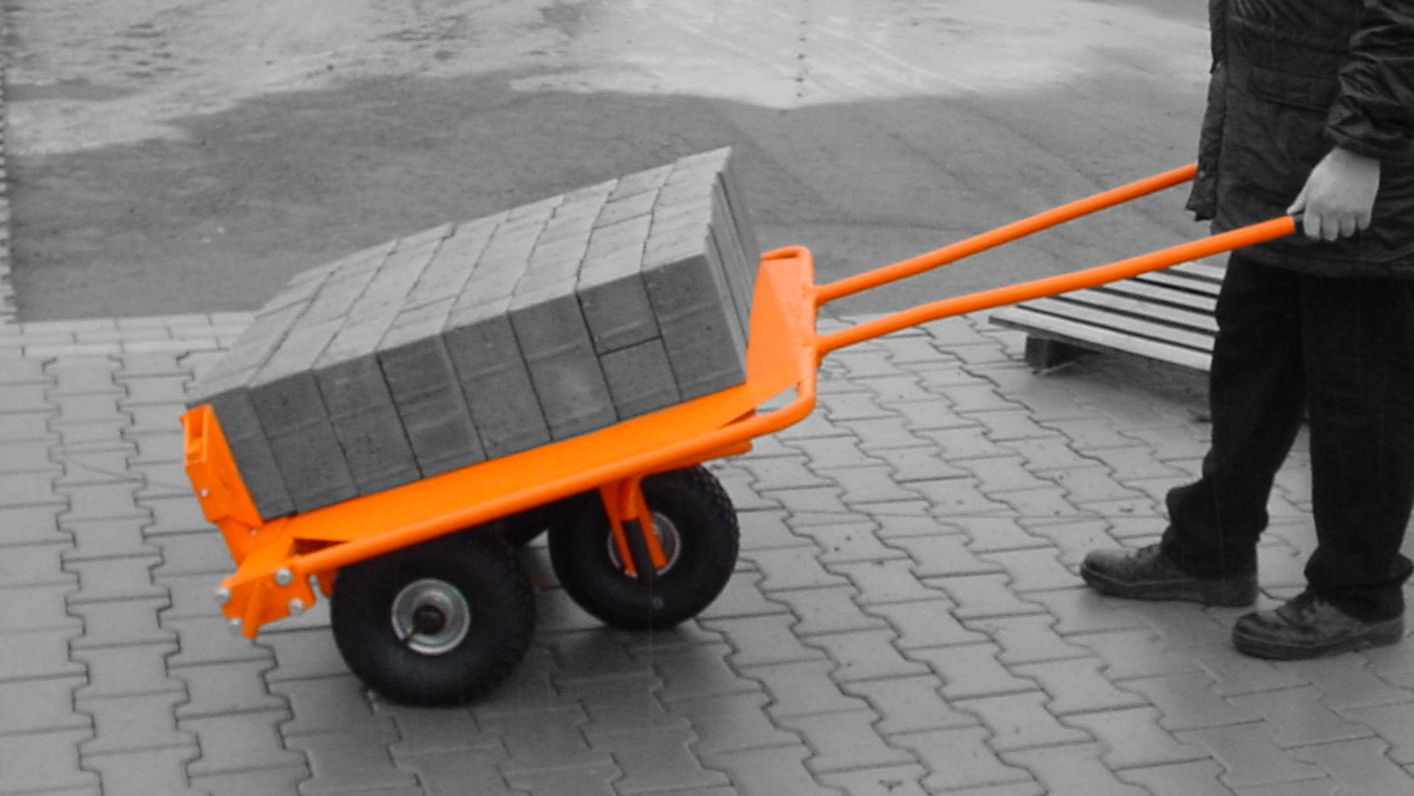 A person pushing an orange wheelbarrow filled with bricks on a paved surface – Optimas.