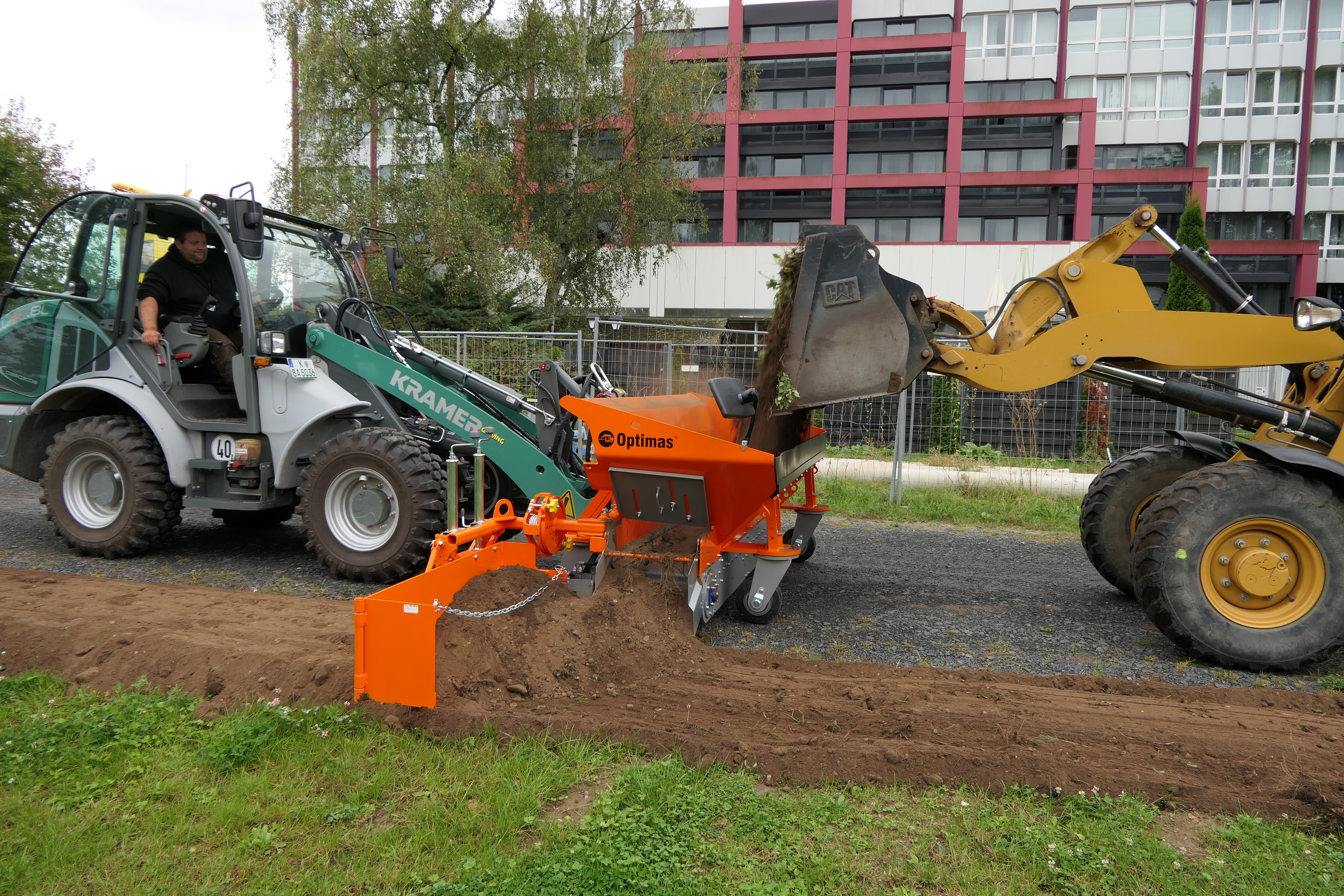 Zwei Lader im Einsatz: ein Kramer-Lader mit einem Mann und ein gelber CAT-Lader beim Erdgraben. Das orangefarbene Optimas-Anbaugerät bearbeitet den Boden effizient – ​​Optimas.