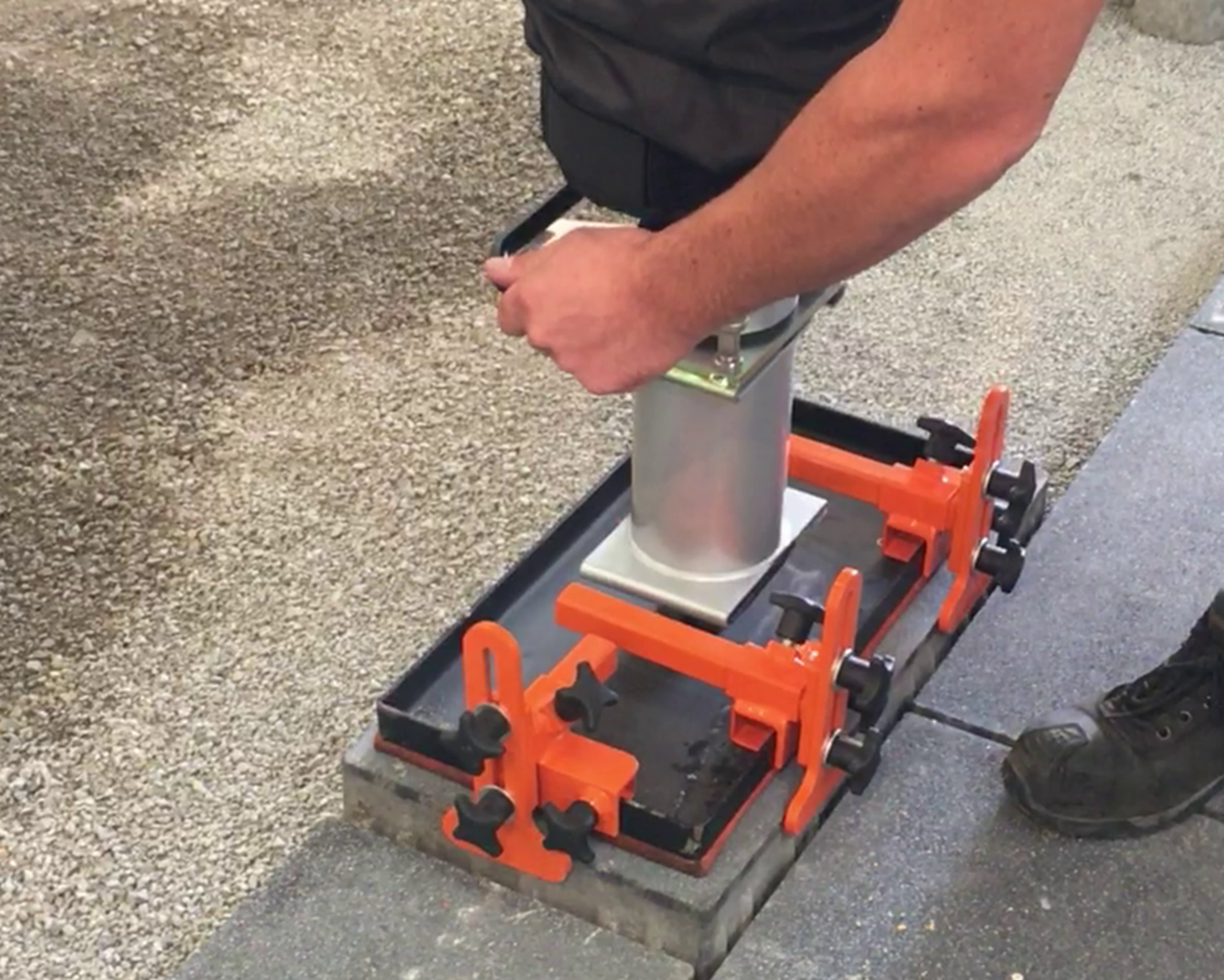 A person adjusts a construction tool on a work surface with orange clamps and a metal base – Optimas.