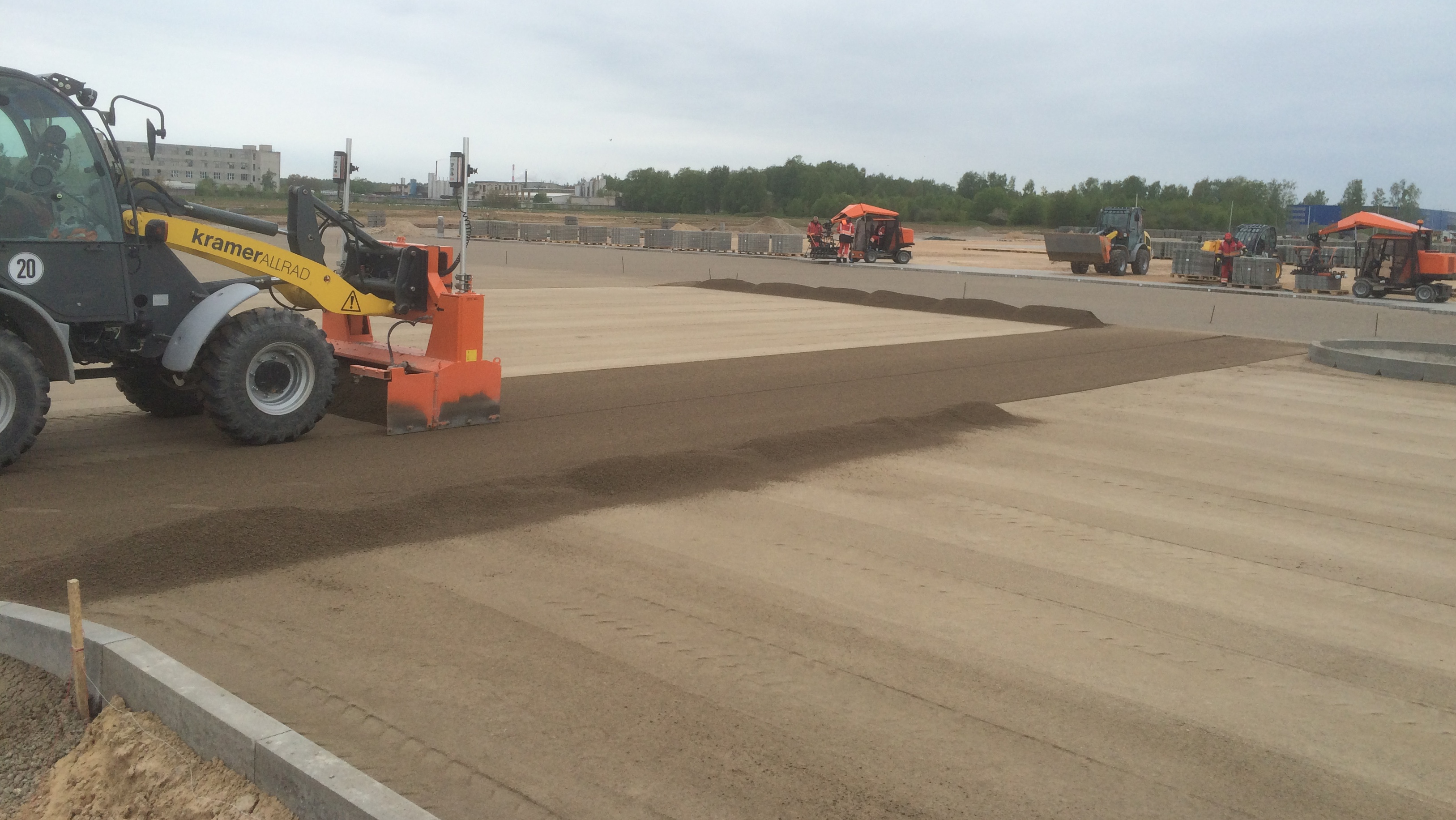 A construction site featuring a Kramer allrad machine leveling sand on a large area, with workers and equipment in the background – Optimas.