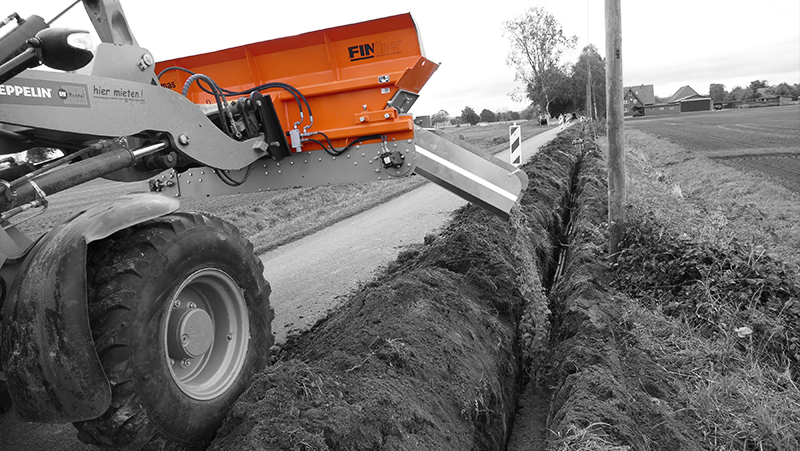 An orange Fin series machine digging a trench beside a rural road, with a focus on the trenching implement and resulting ditch – Optimas