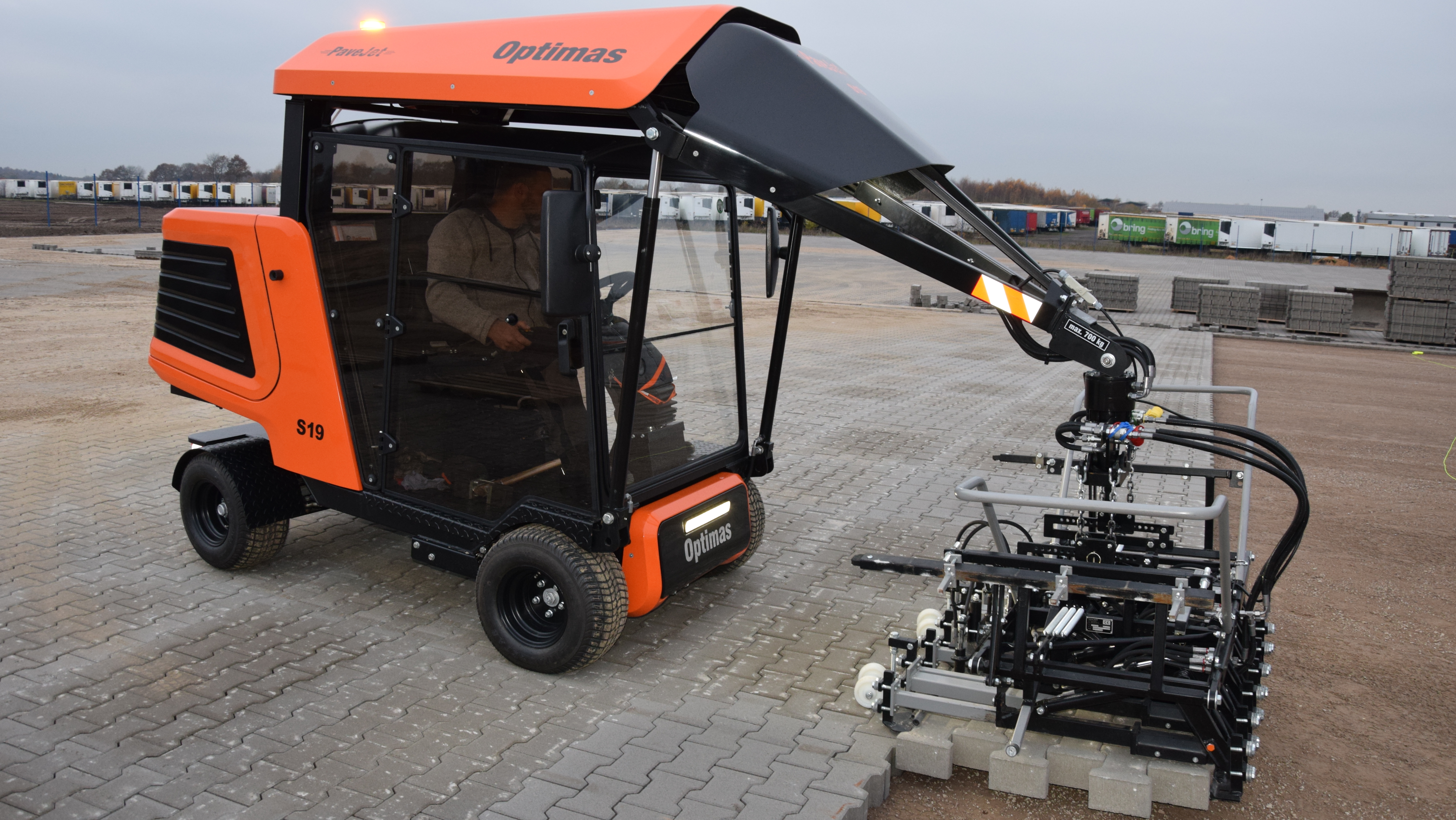 An orange and black compact machine with a crane arm is operating on a construction site, handling materials – Optimas.