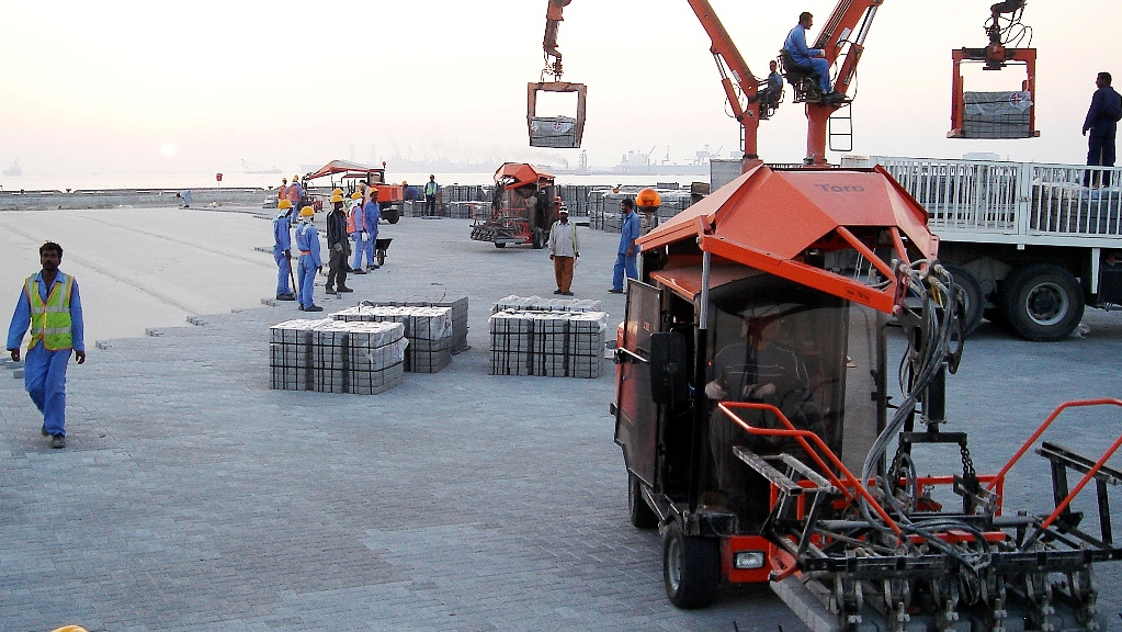 Workers operating machinery and handling materials at a construction site near the waterfront during sunset – Optimas.