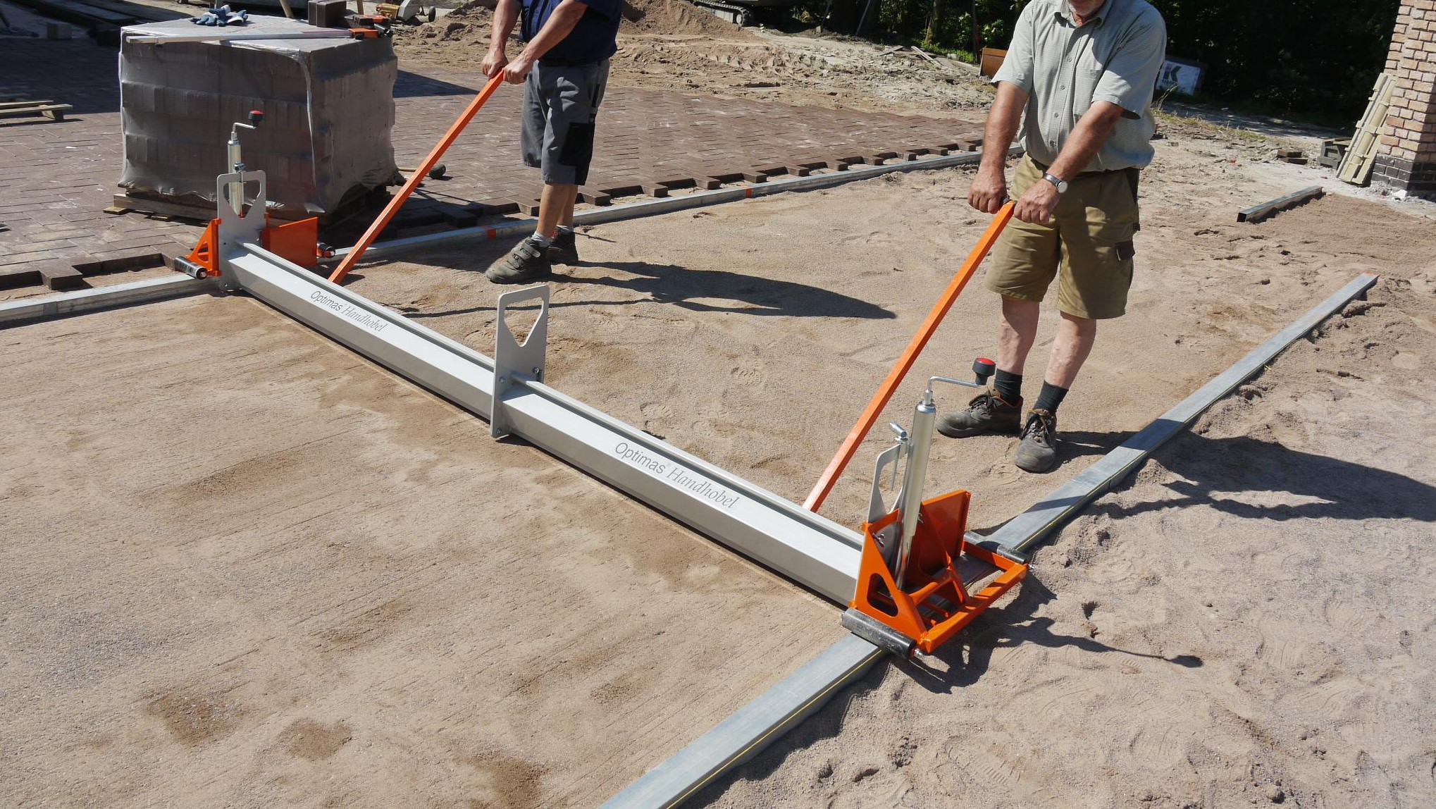 Two workers leveling sand using a large screed tool on a construction site for paving. – Optimas