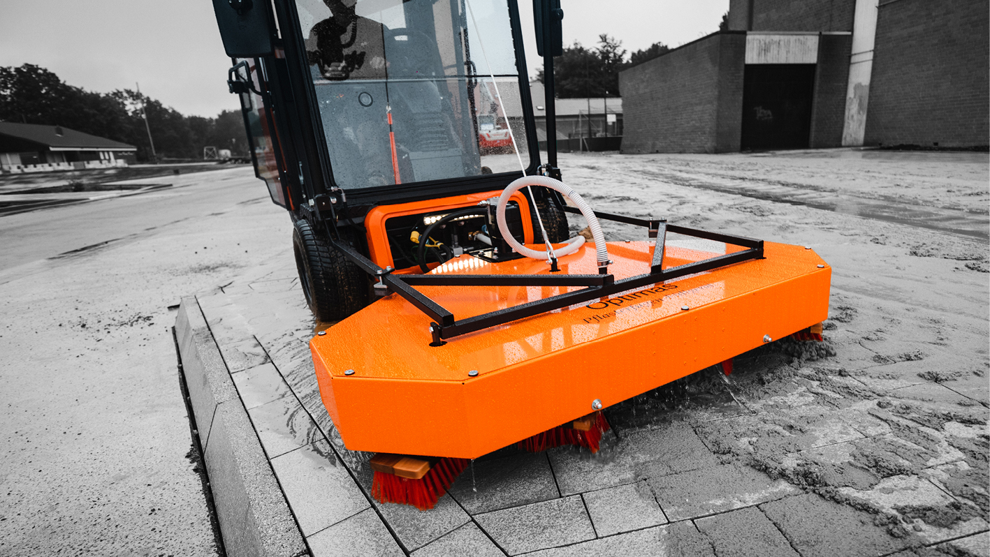 Close-up of an orange Optimas Pflasterreiniger road cleaning machine on a wet, grey paved road with buildings in the background – Optimas.