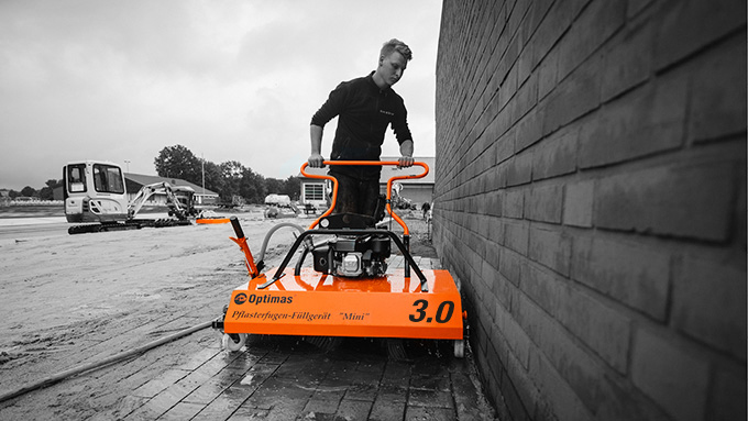 A worker operates a wall plastering machine next to a brick wall at a construction site – Optimas.