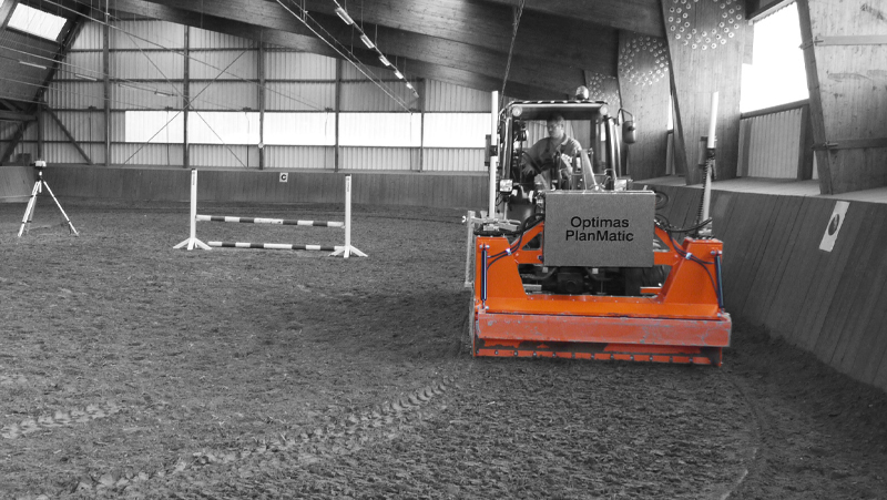 Orange arena leveller machine, an Optimas PlanMatic, operated indoors, prepares the sand surface near a hurdle with a person driving – Optimas.