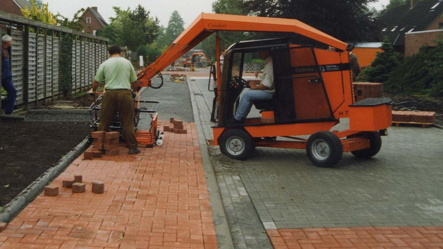 A worker operates a brick-laying machine on a newly paved road, while another worker assists nearby – Optimas.