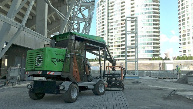 A green construction vehicle operates on a building site with modern skyscrapers in the background – Optimas.