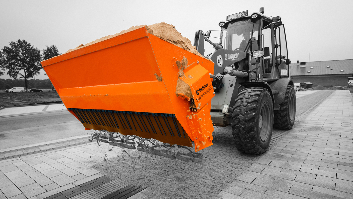 A construction vehicle with an orange bucket is unloading sand onto a paved surface – Optimas.