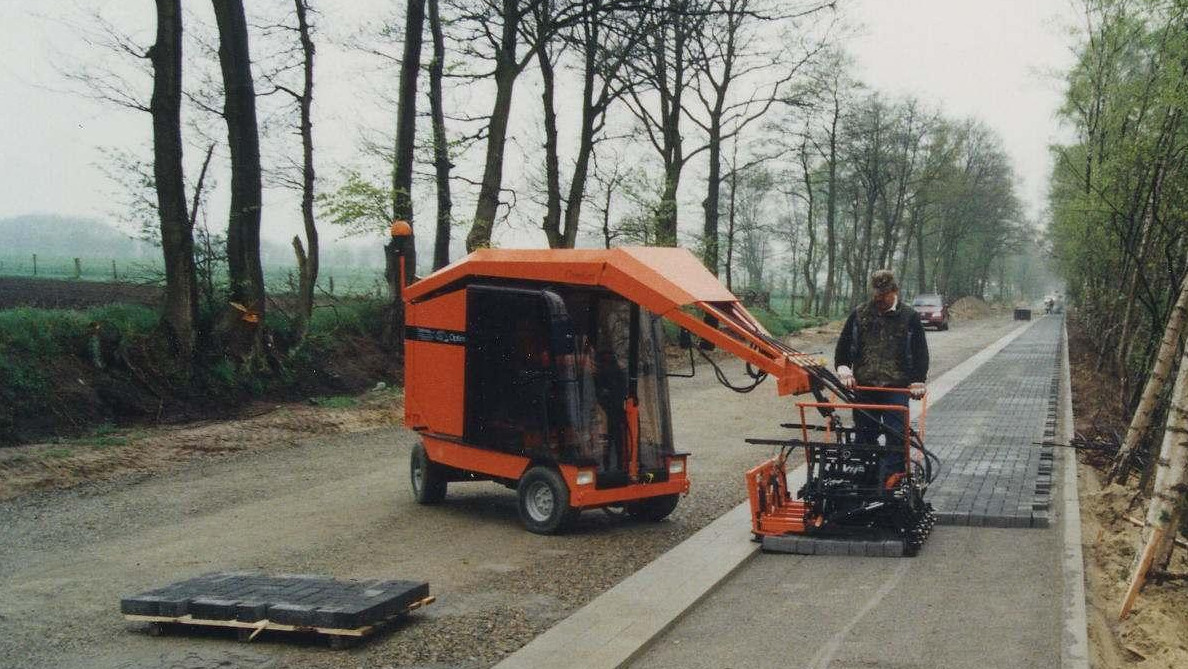 A worker operates an orange paving machine on a gravel road, laying down paving stones alongside trees – Optimas.