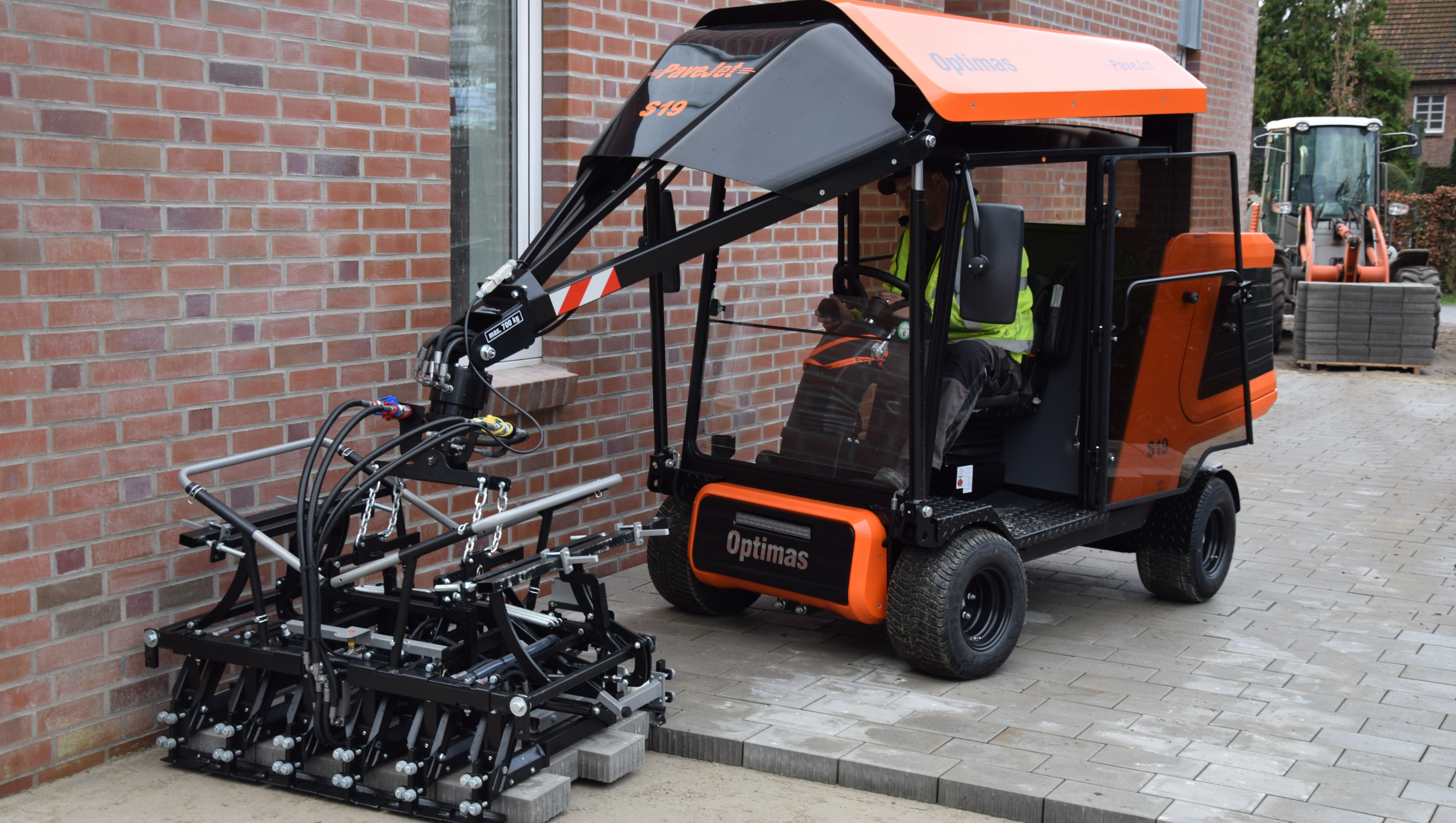 A compact utility vehicle with an attached implement, parked on a paved surface near a brick wall – Optimas.