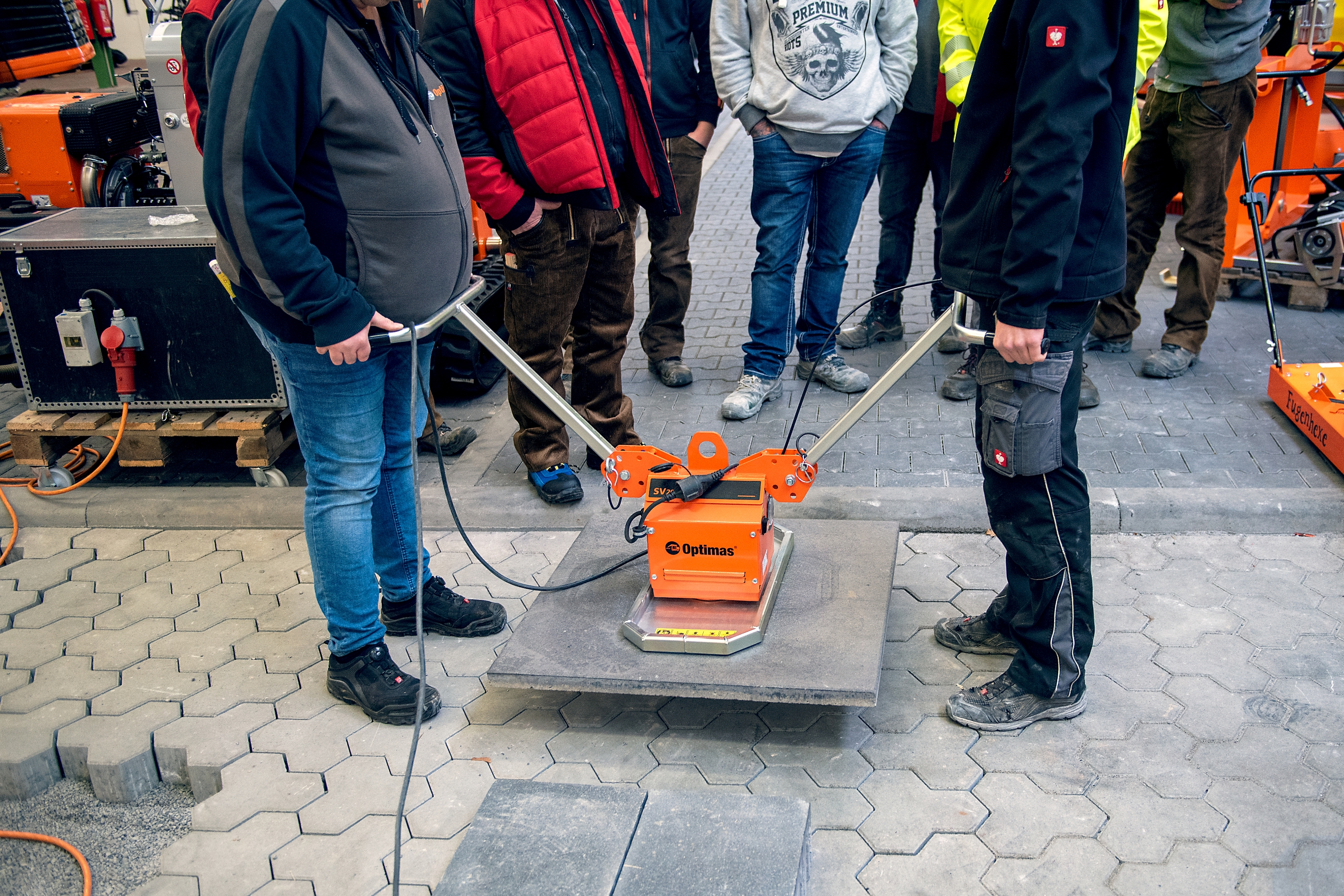 Zwei Arbeiter führen eine Pflastermaschine vor, während eine Gruppe in einer Werkstatt zuschaut – Optimas.