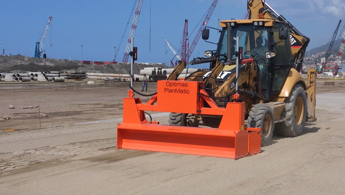 A construction vehicle with an orange attachment is working on a site with cranes and pipes in the background – Optimas.