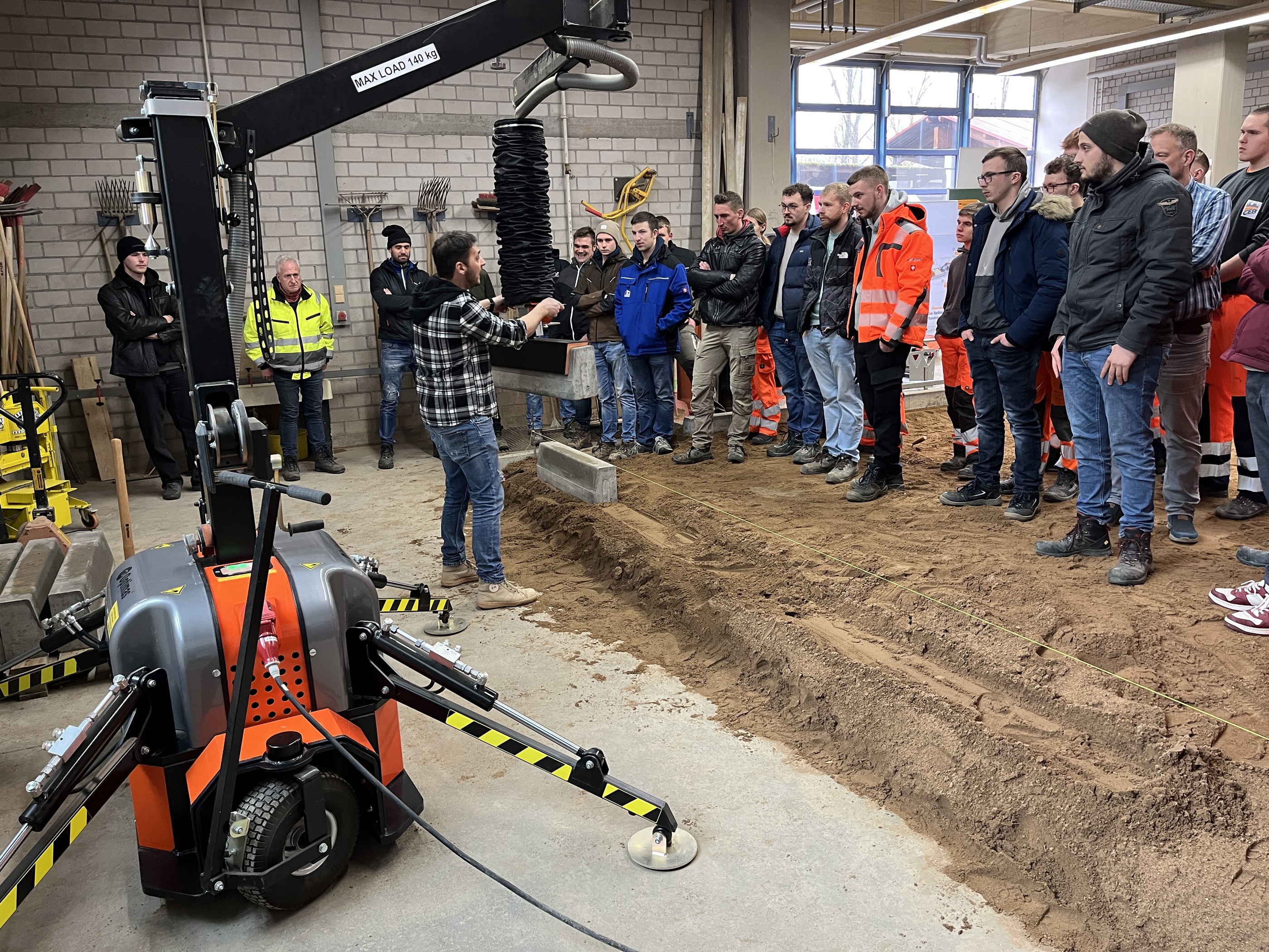 Eine Gruppe von Bauarbeitern beobachtet eine Demonstration der Optimas-Vakuumhebemaschine, die zum Verlegen von Pflastersteinen entlang einer Grabenlinie verwendet wird.