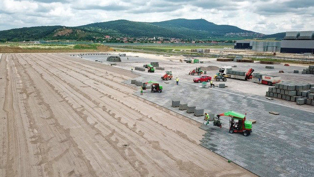 Construction workers laying paving stones on a large site with machinery and hills in the background – Optimas.