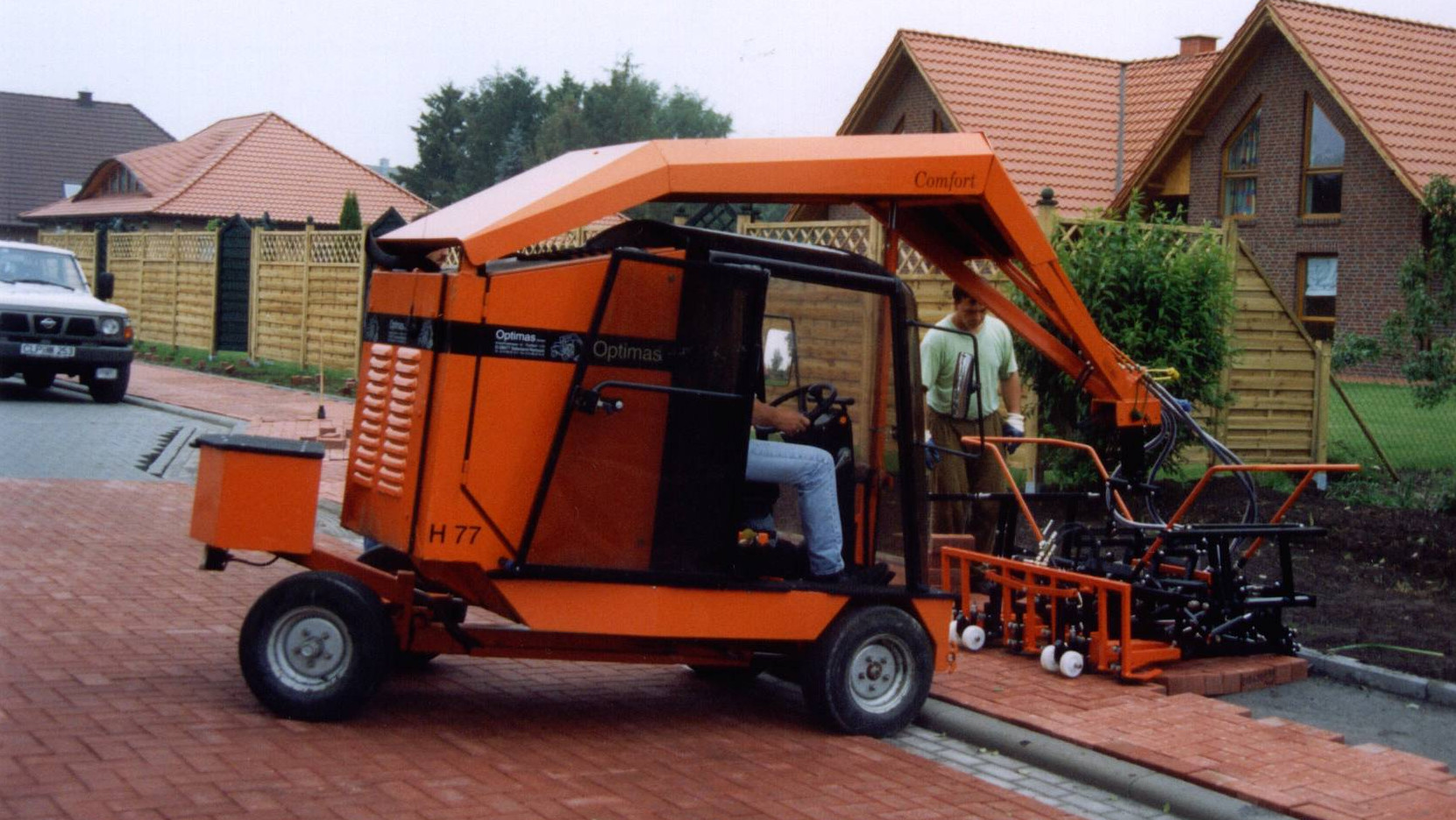 A brick-laying machine in operation, with a worker guiding the process on a residential street – Optimas.