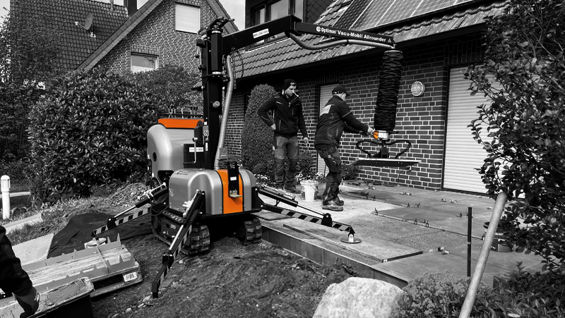 Two construction workers use an Optimas Vacu-Mobil Allrounder to install stone slabs outside a home, streamlining paving projects – Optimas.
