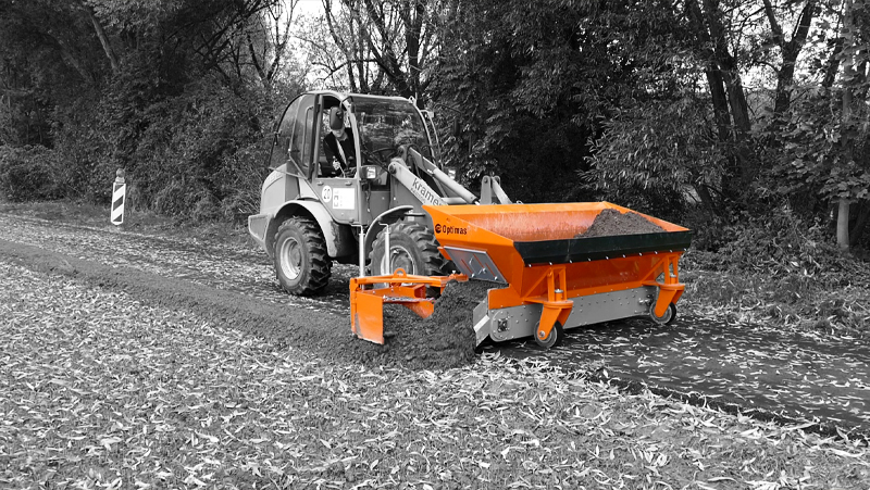 Image displays a Kramer tractor in a monochrome landscape, fitted with a vibrant orange Optimas attachment spreading material on a rural path – Optimas.