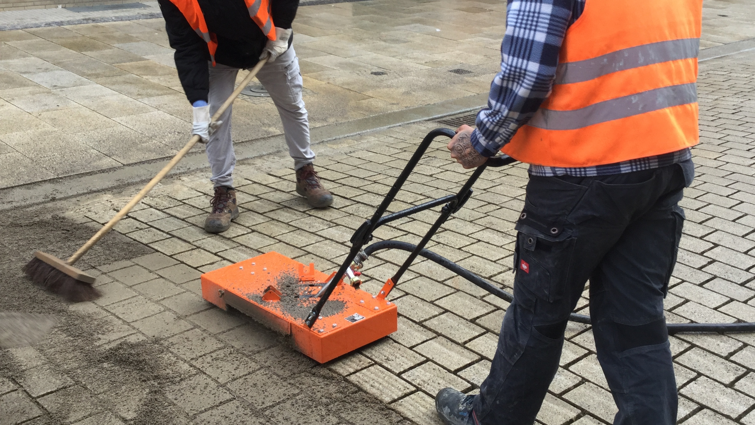 Two construction workers in safety vests using a machine to clean and level pavement stones – Optimas.