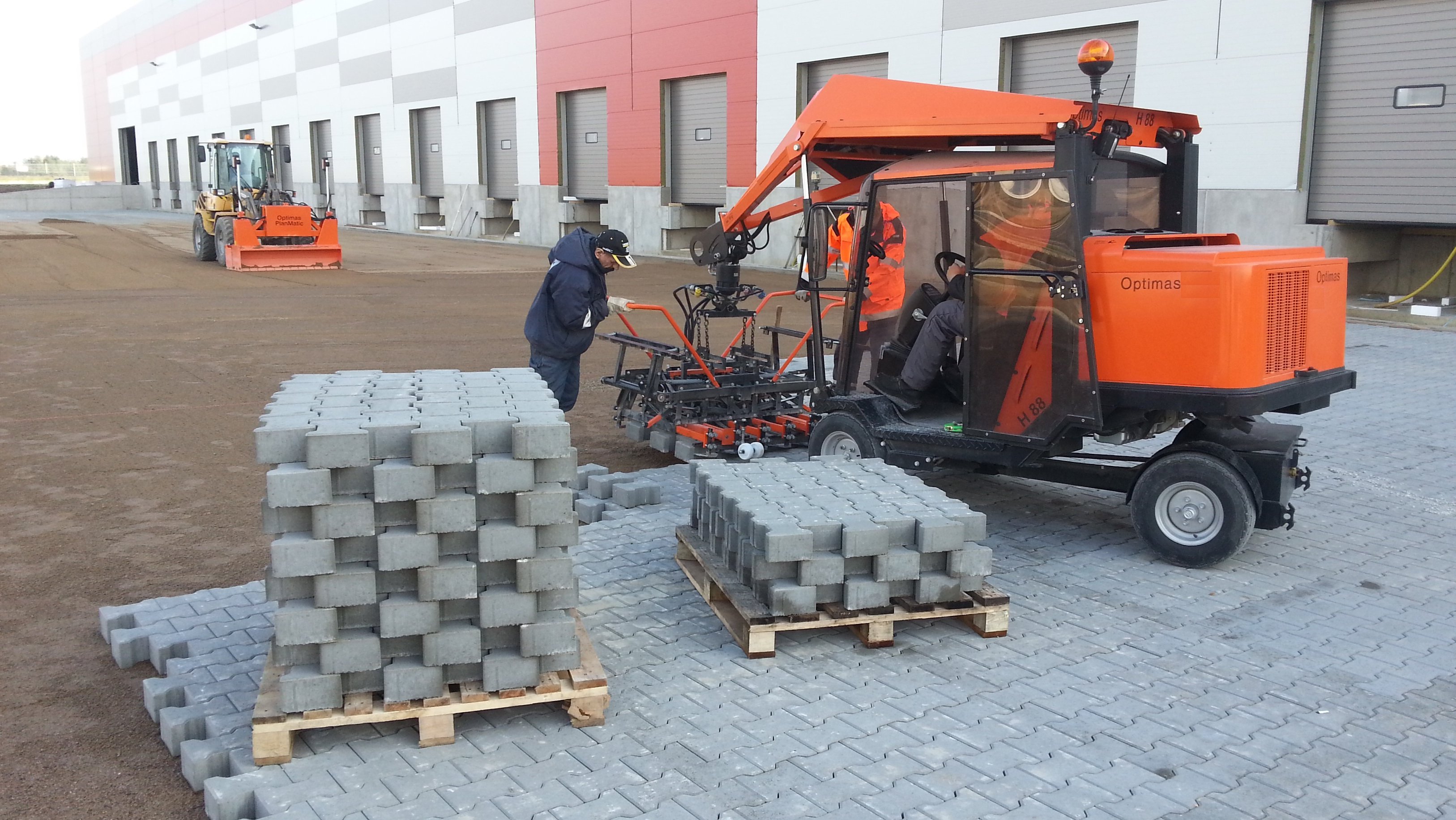 A construction site with workers using machinery to lay paving stones on a gravel surface – Optimas.