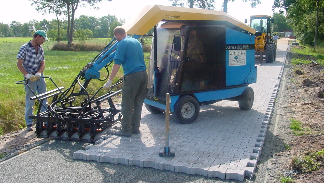 Two workers laying paving stones using a specialized machine on a grassy area, with construction equipment in the background – Optimas.