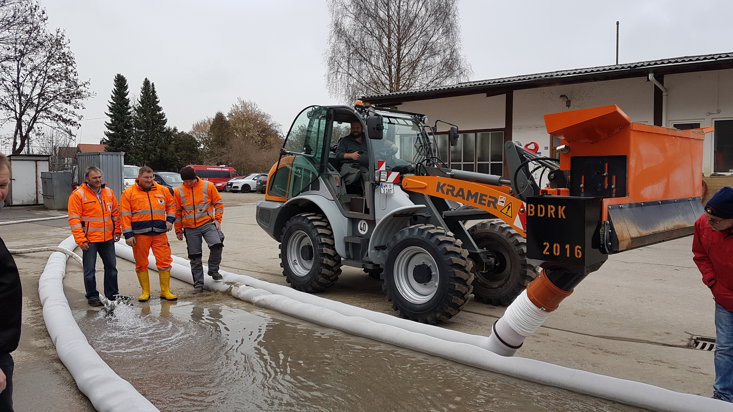 A construction site with workers in orange uniforms and a Kramer loader managing water with a hose – Optimas.