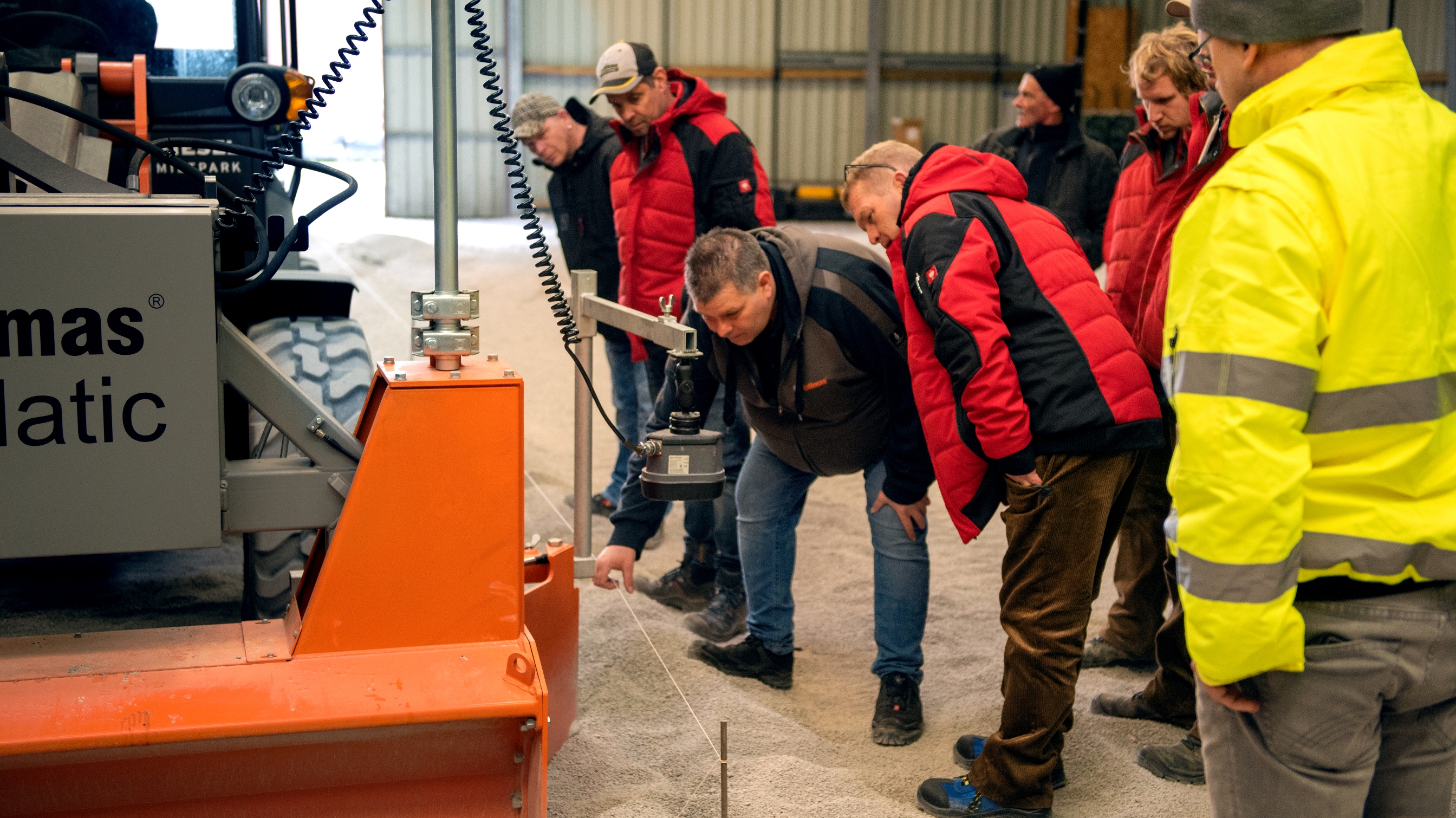 Eine Gruppe von Menschen beobachtet die Vorführung einer Baumaschine in einer Lagerhalle – Optimas.