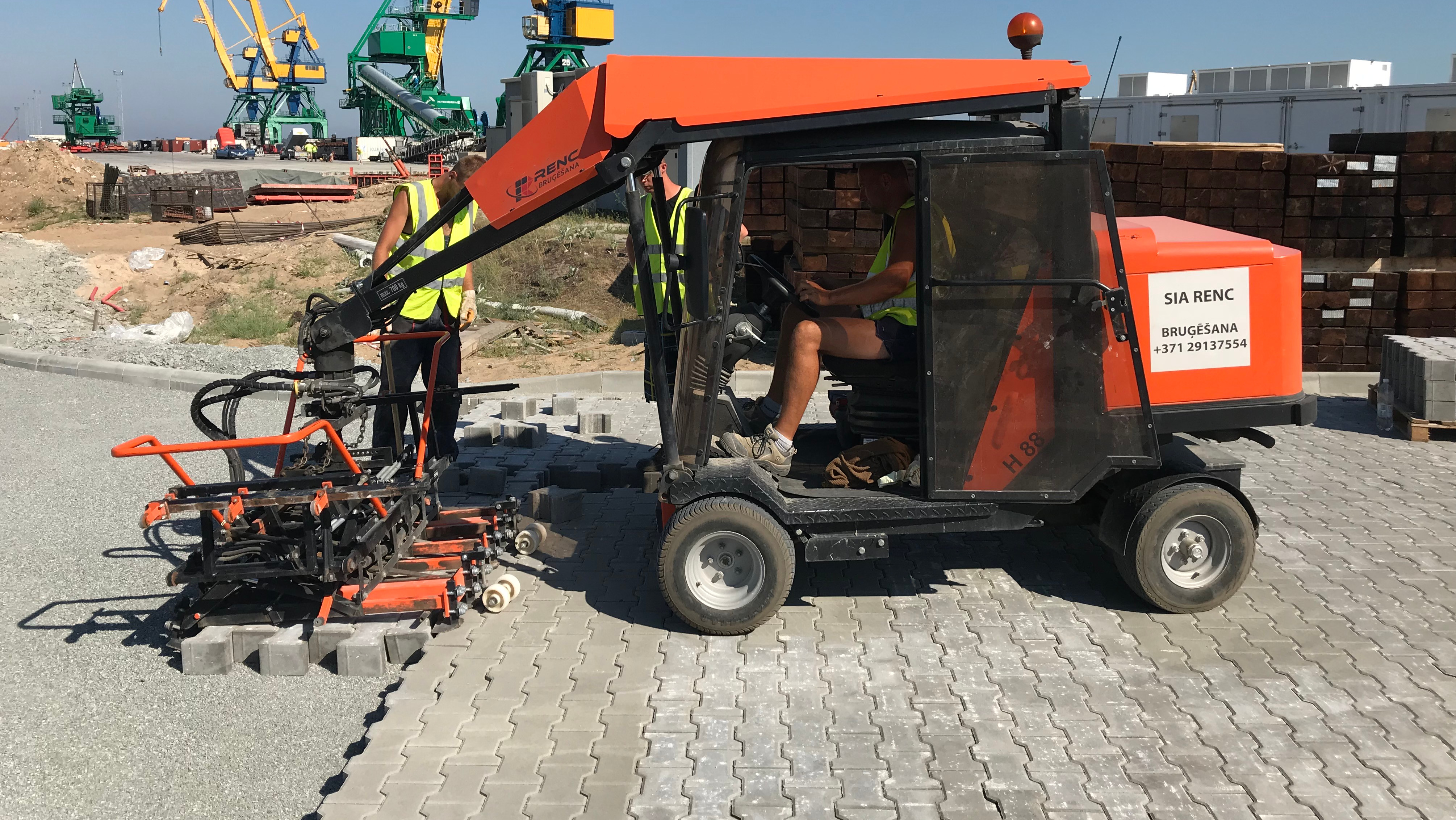 A small construction vehicle laying paving stones with workers in safety vests on a construction site – Optimas.