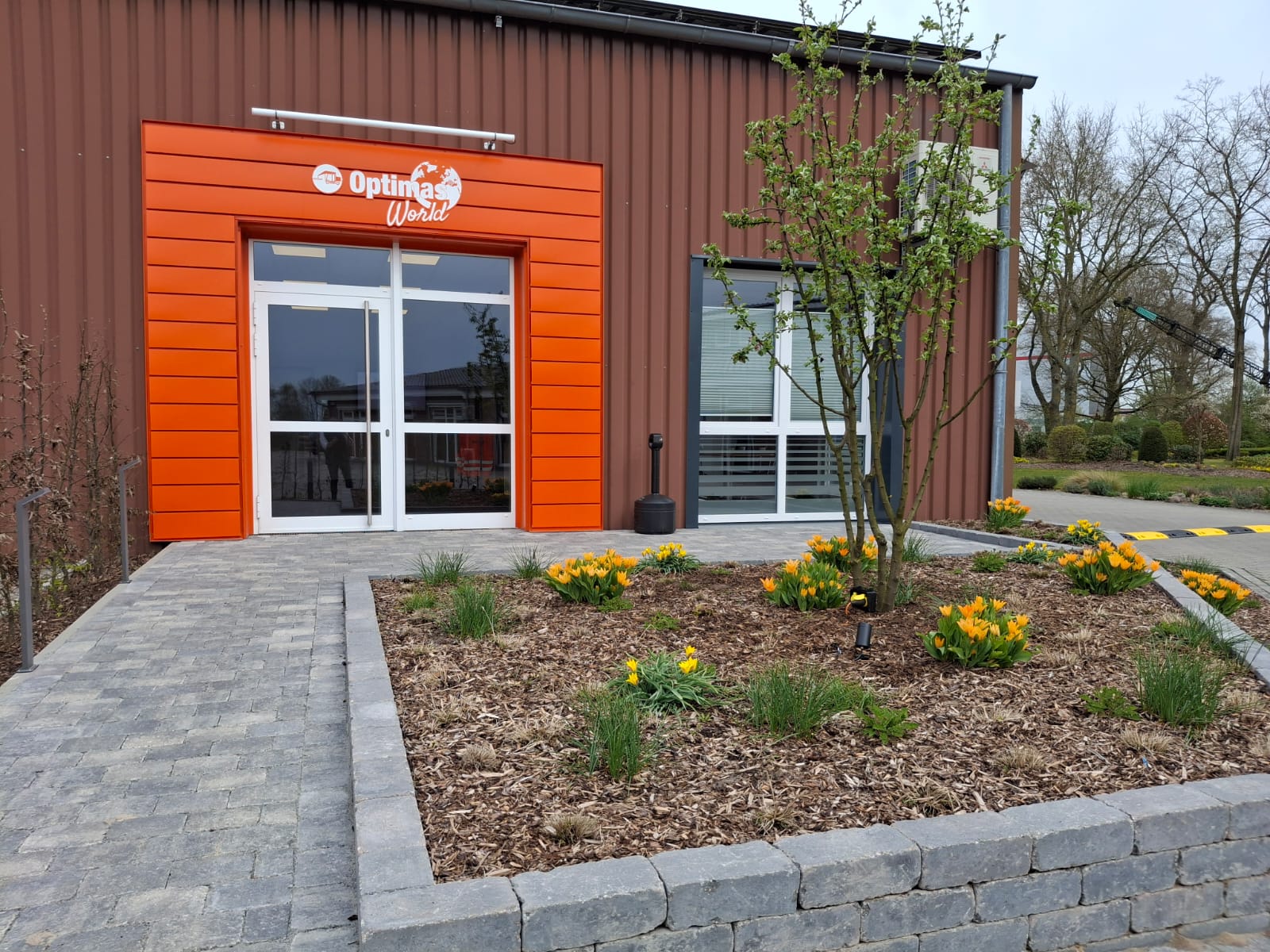 Entrance of Optimas World featuring a vibrant orange facade and landscaped flower beds – Optimas.