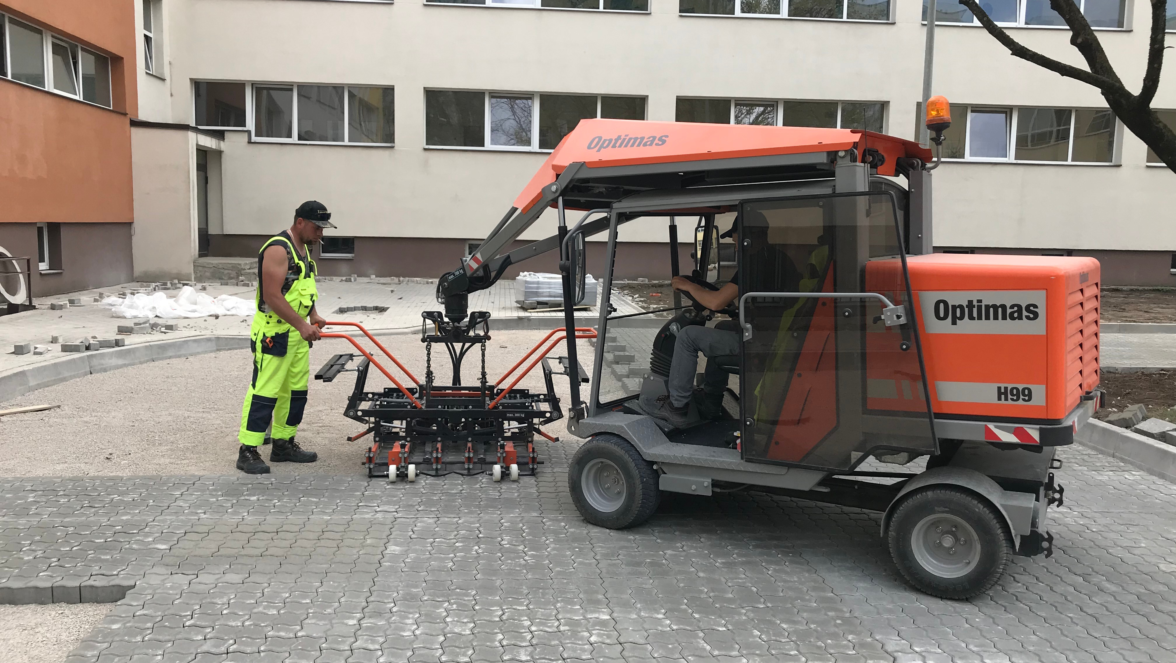 A construction worker in a bright vest operates a compact machine for paving stones in a work area – Optimas.