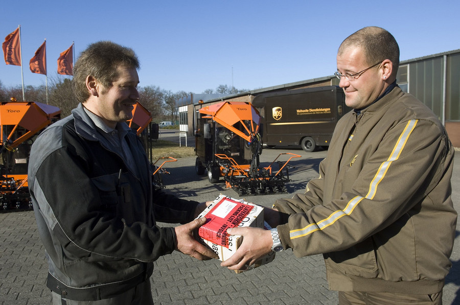 Two men exchanging a package outdoors with delivery vehicles in the background – Optimas.