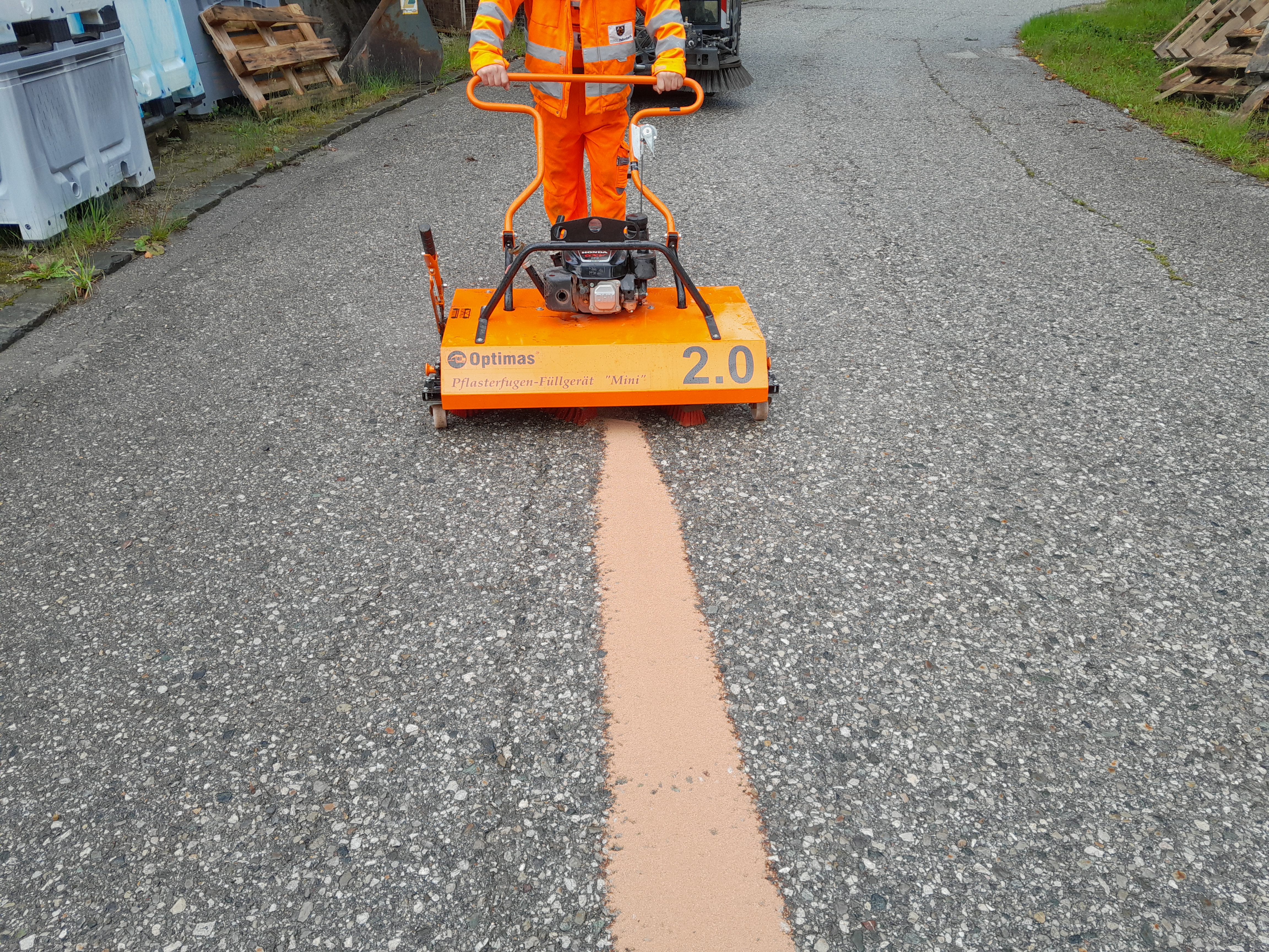 A worker operates an orange filling machine on a gravel road, applying a light brown material – Optimas.