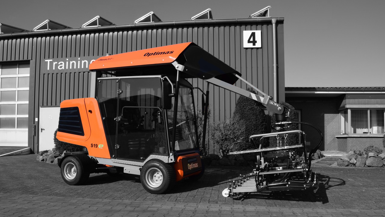 A compact, orange and gray machine with a lifting arm parked outside a training facility – Optimas.