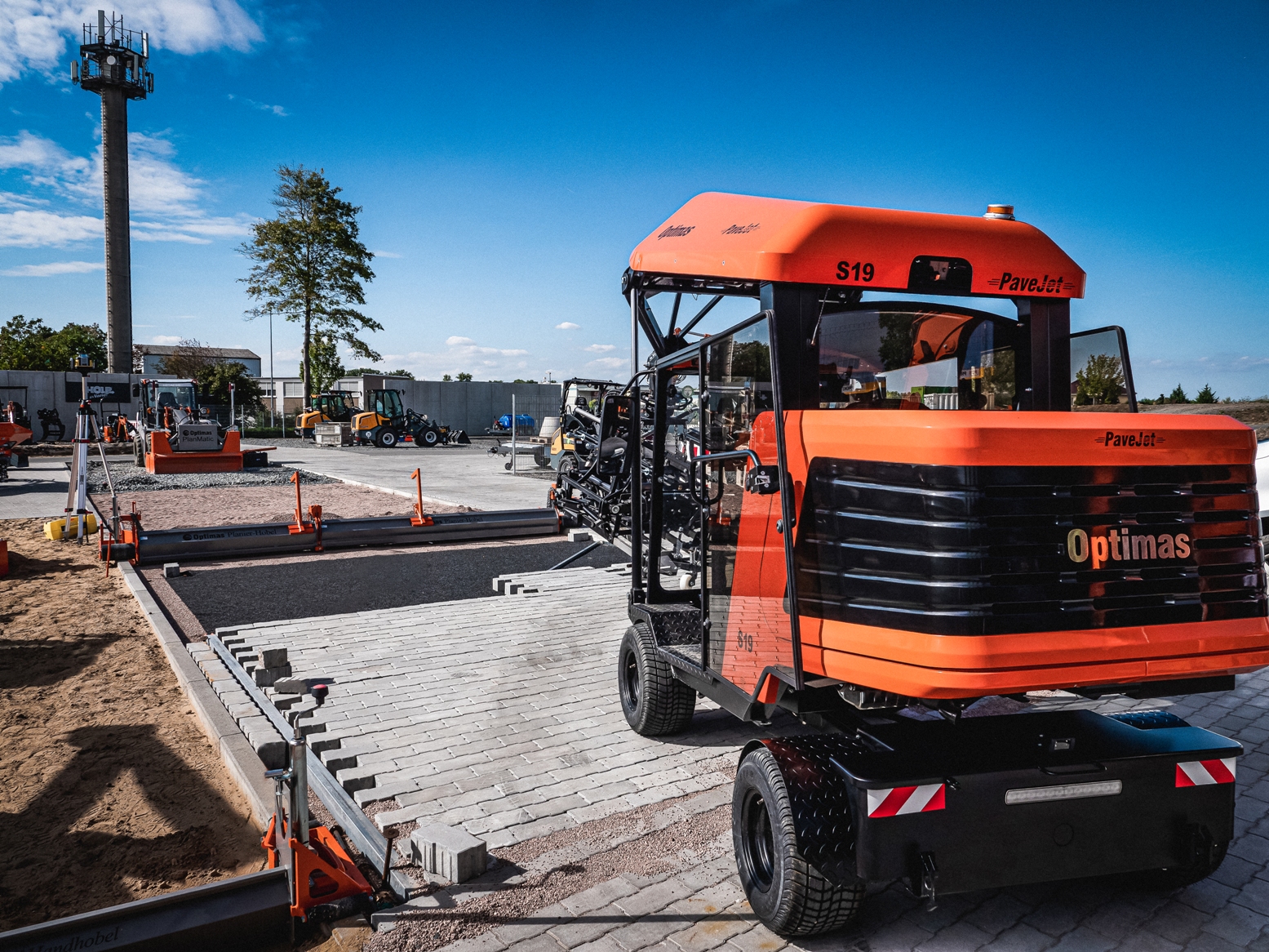 A PaveJet S19 machine from Optimas is paving a surface at a construction site under a clear blue sky – Optimas.