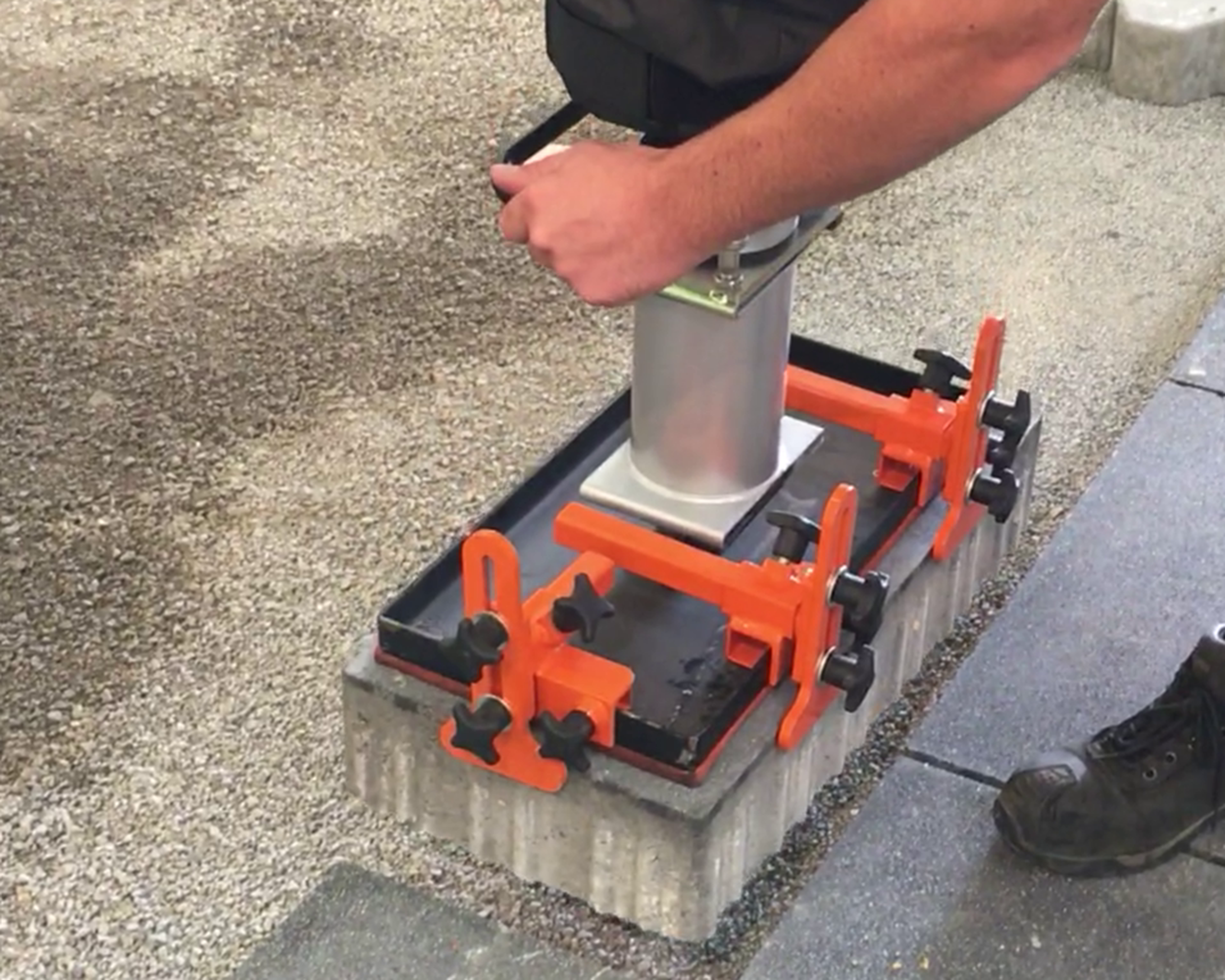 A worker using a tool to secure a metal post on a concrete base with orange clamps on gravel surface – Optimas.