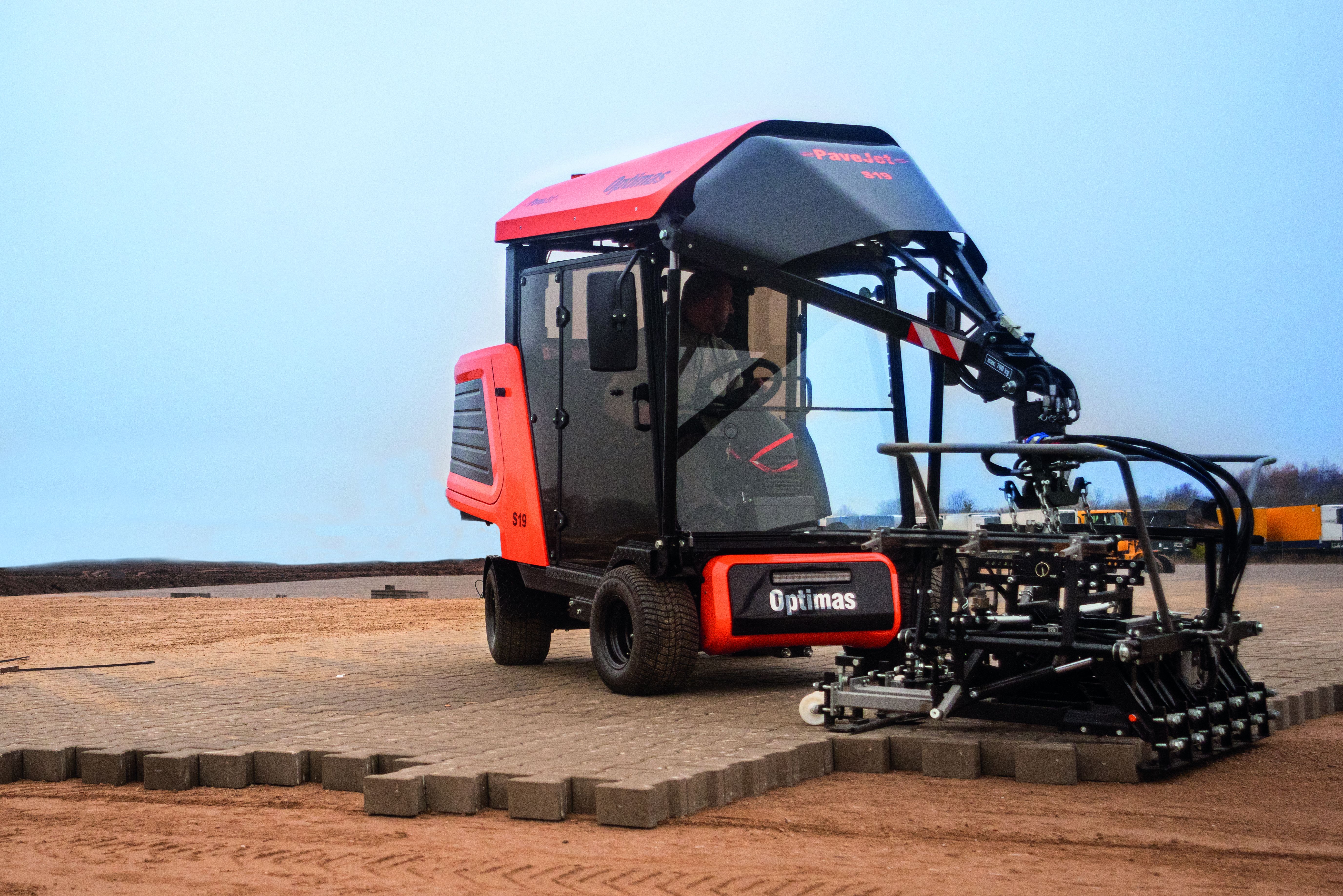 A compact paving machine with a red and black design is laying bricks on a construction site – Optimas.