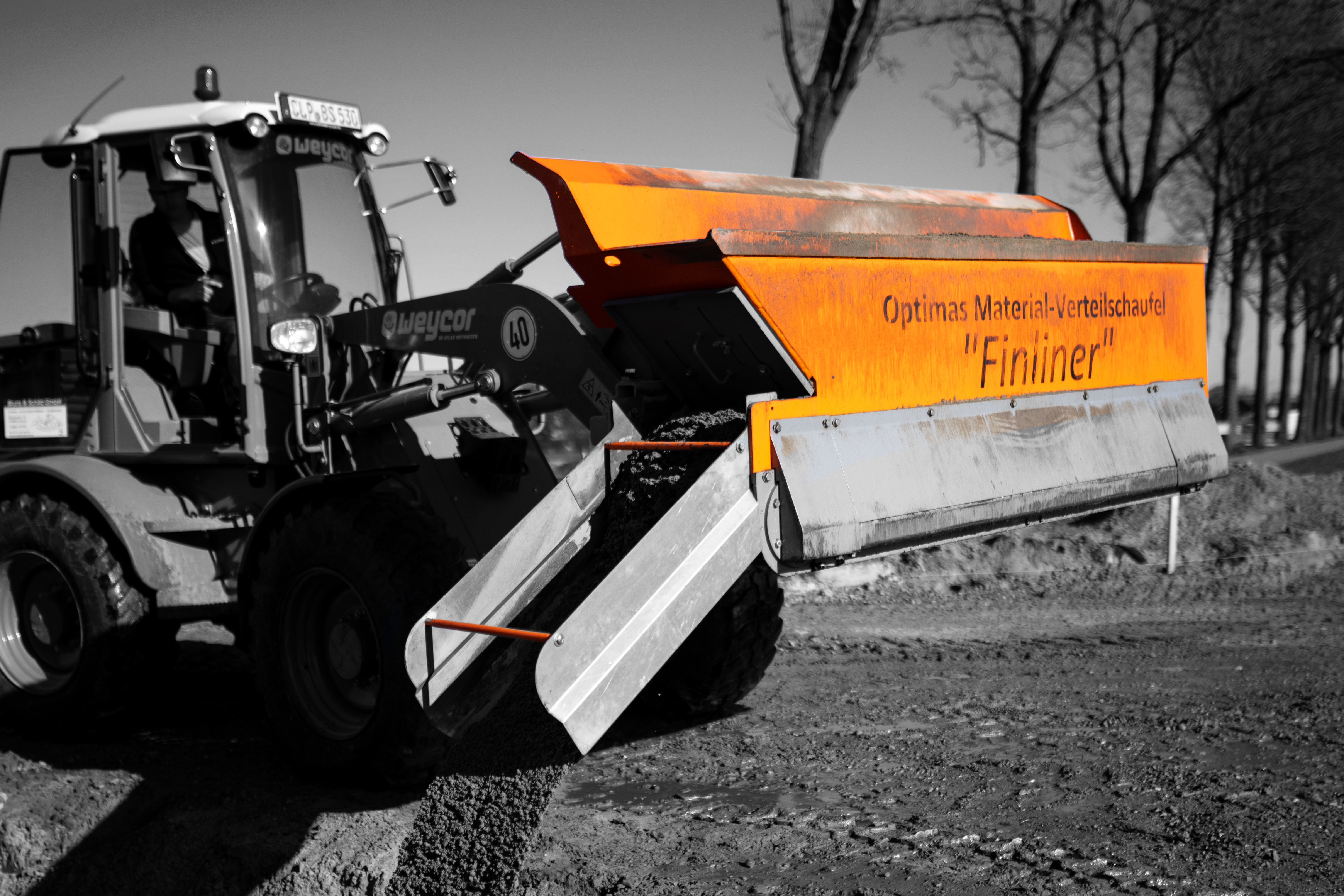 A construction vehicle with an orange material distribution shovel labeled 'Finliner' in action on a dirt road – Optimas.