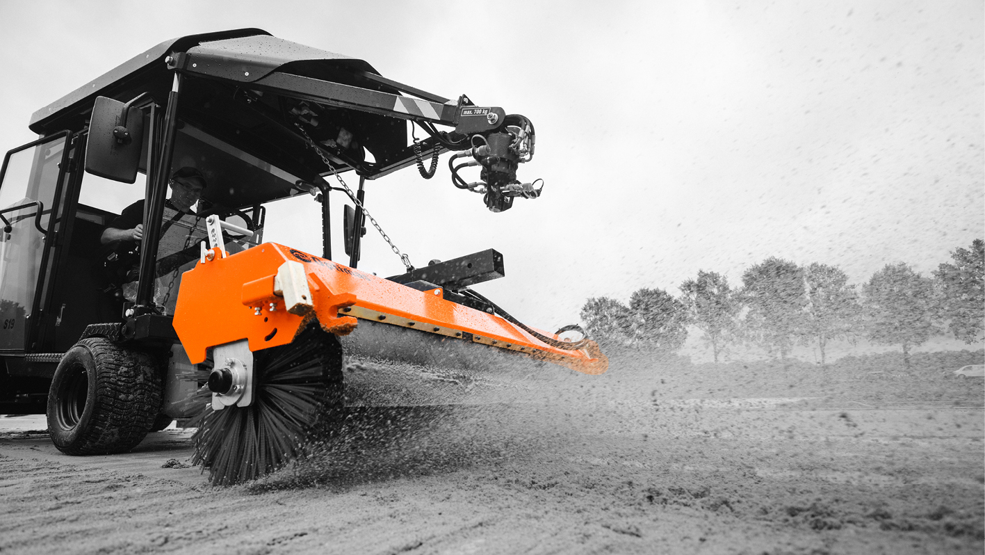 A worker operates a machine with an orange broom attachment, clearing sand while dust clouds rise – Optimas.
