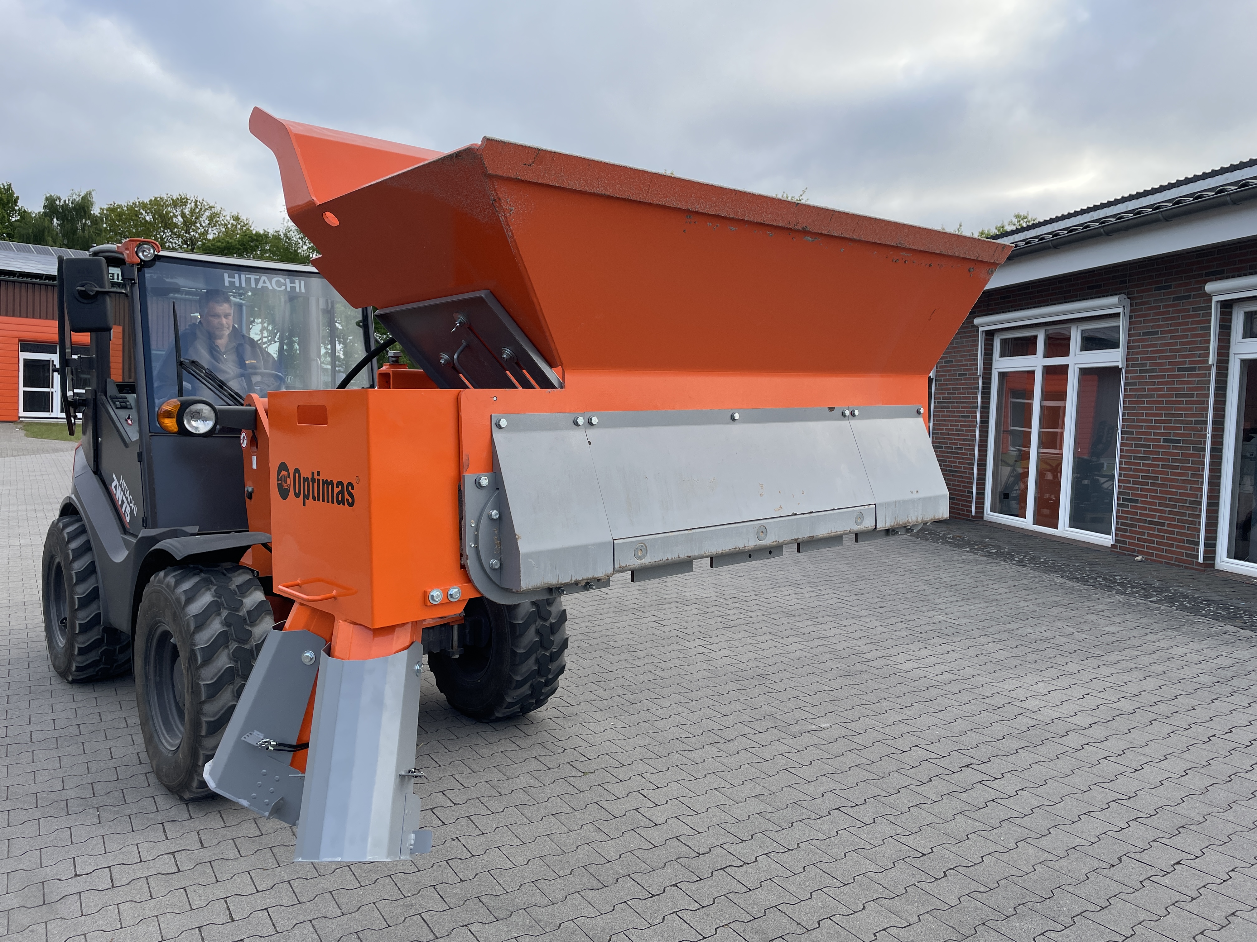 A Hitachi loader with an orange Optimas attachment, parked on a paved area outside a building – Optimas.