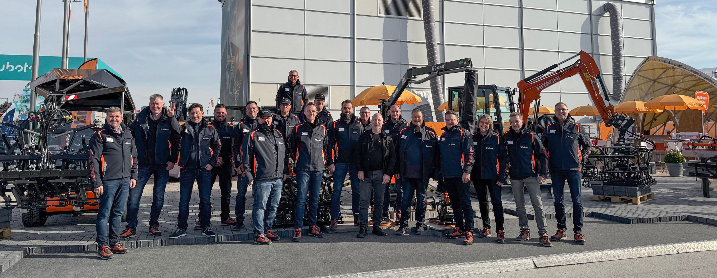 Group photo of Optimas employees standing in front of construction equipment, showcasing a team unified in the construction industry – Optimas.