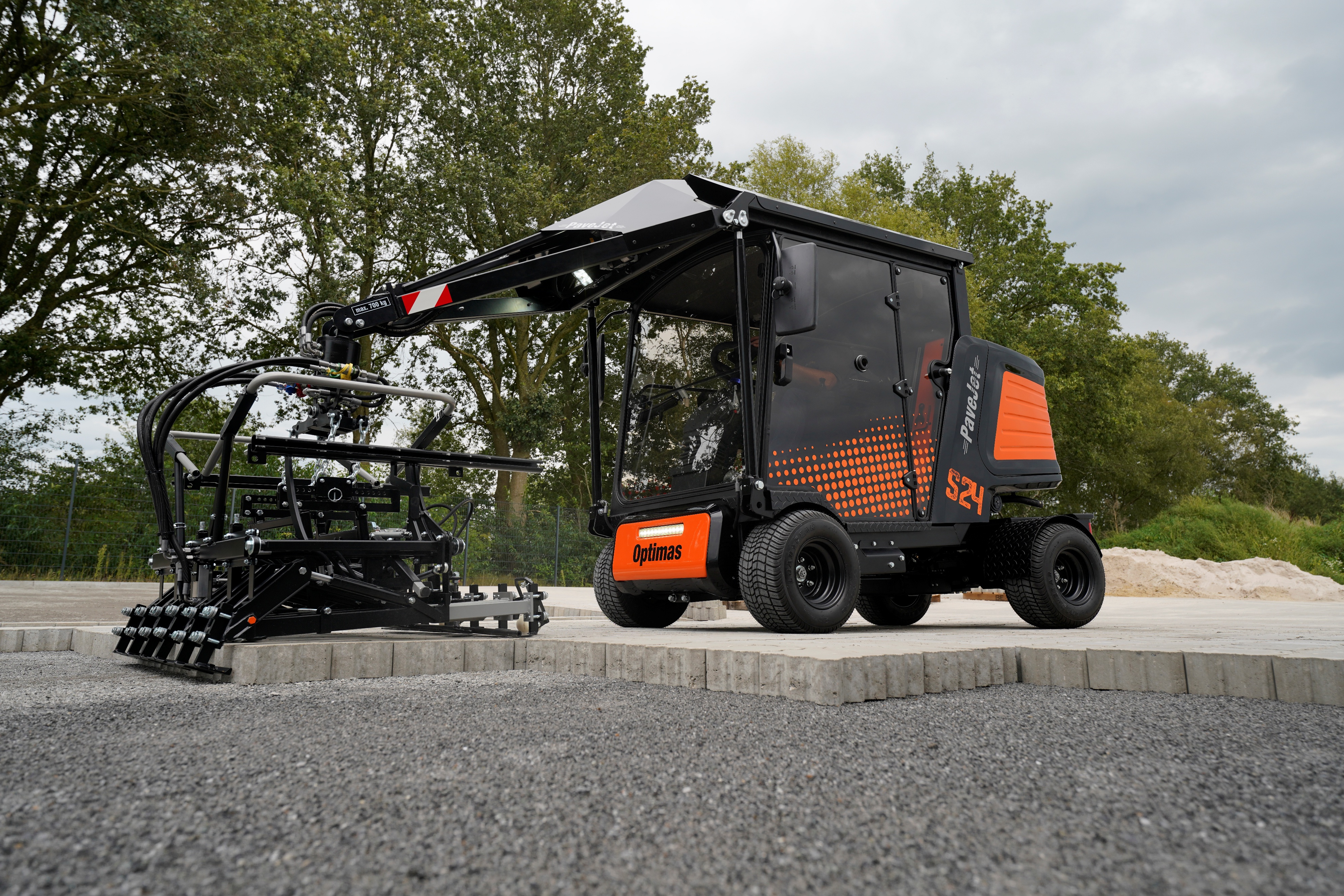 Die Pflastermaschine Optimas Paverjet S24, schwarz und orange, geparkt auf Kies und Beton, mit Bäumen im Hintergrund, präsentiert ihr Design – Optimas.