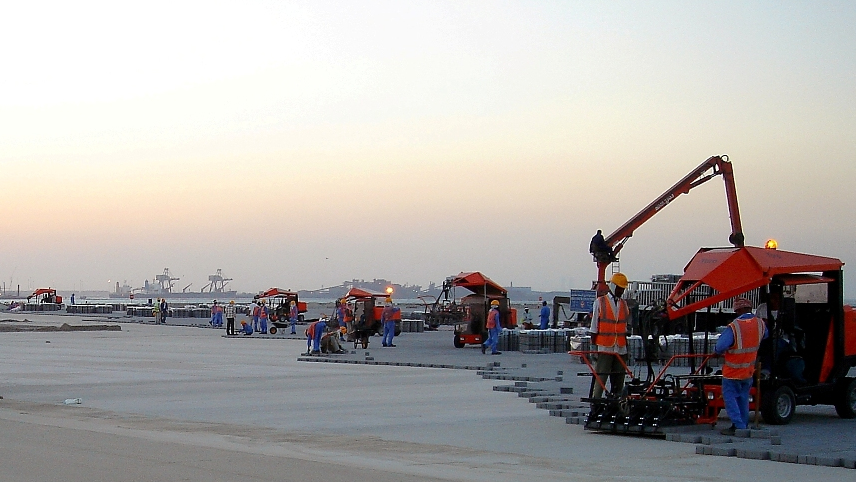 Construction workers and machinery are busy laying pavement at a construction site during sunset – Optimas.