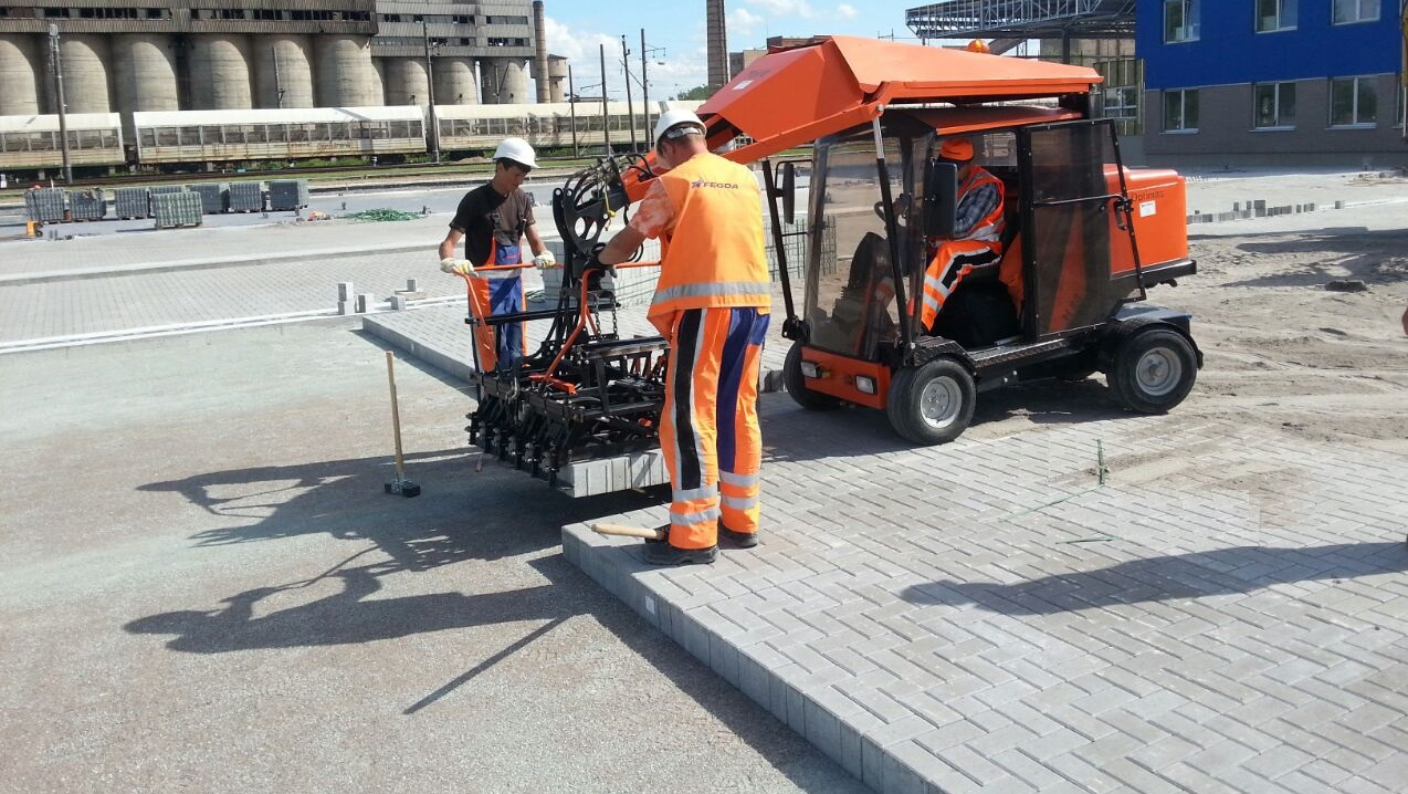 Two workers use a machine to lay bricks on a construction site, with machinery and buildings in the background – Optimas.