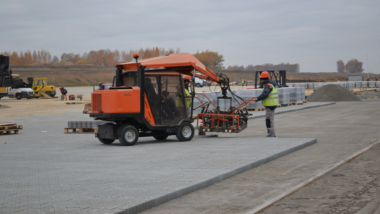 A construction worker operates a small machine laying paving stones on a site, with workers in the background – Optimas.