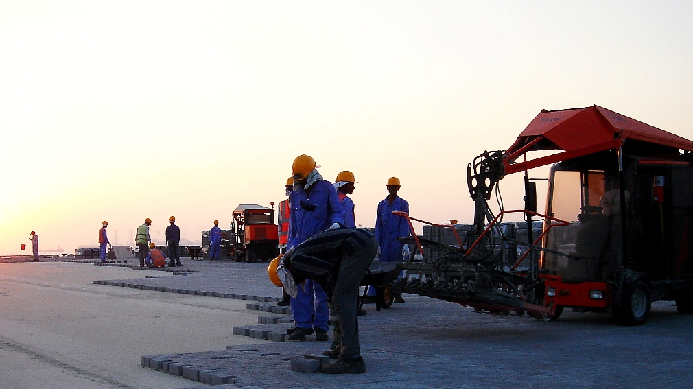 Workers in safety gear laying pavement blocks at a construction site during sunset – Optimas.