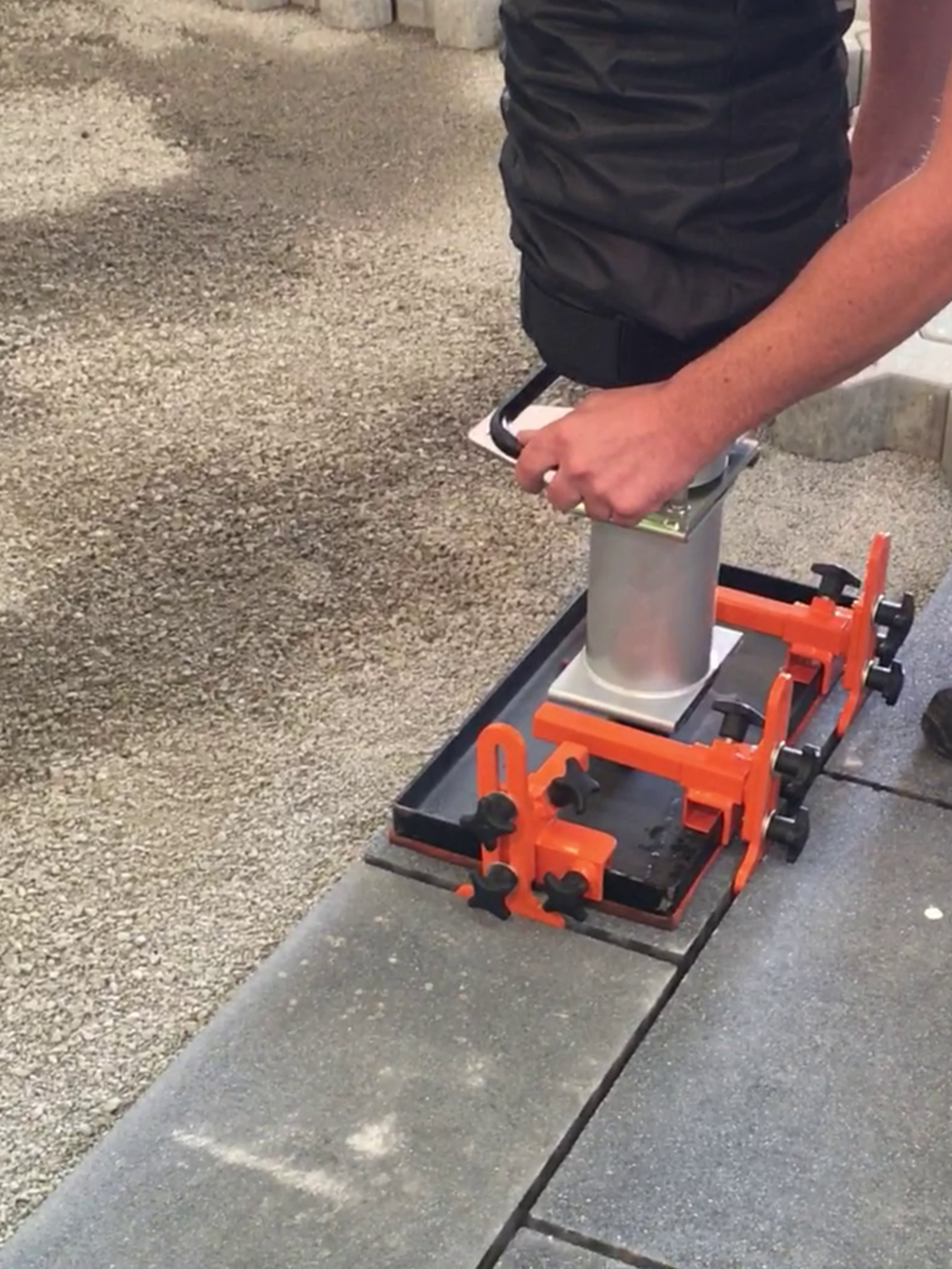 A person using a paving stone lifting tool on a gravel surface to position concrete slabs – Optimas.
