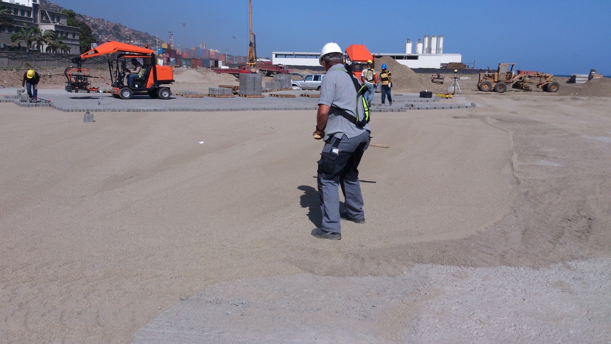 Construction workers operate machinery on a sandy site with containers in the background – Optimas.