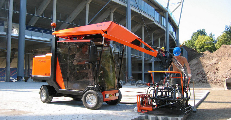 A construction worker operates an orange Toro machine on a building site, with a stadium in the background – Optimas.
